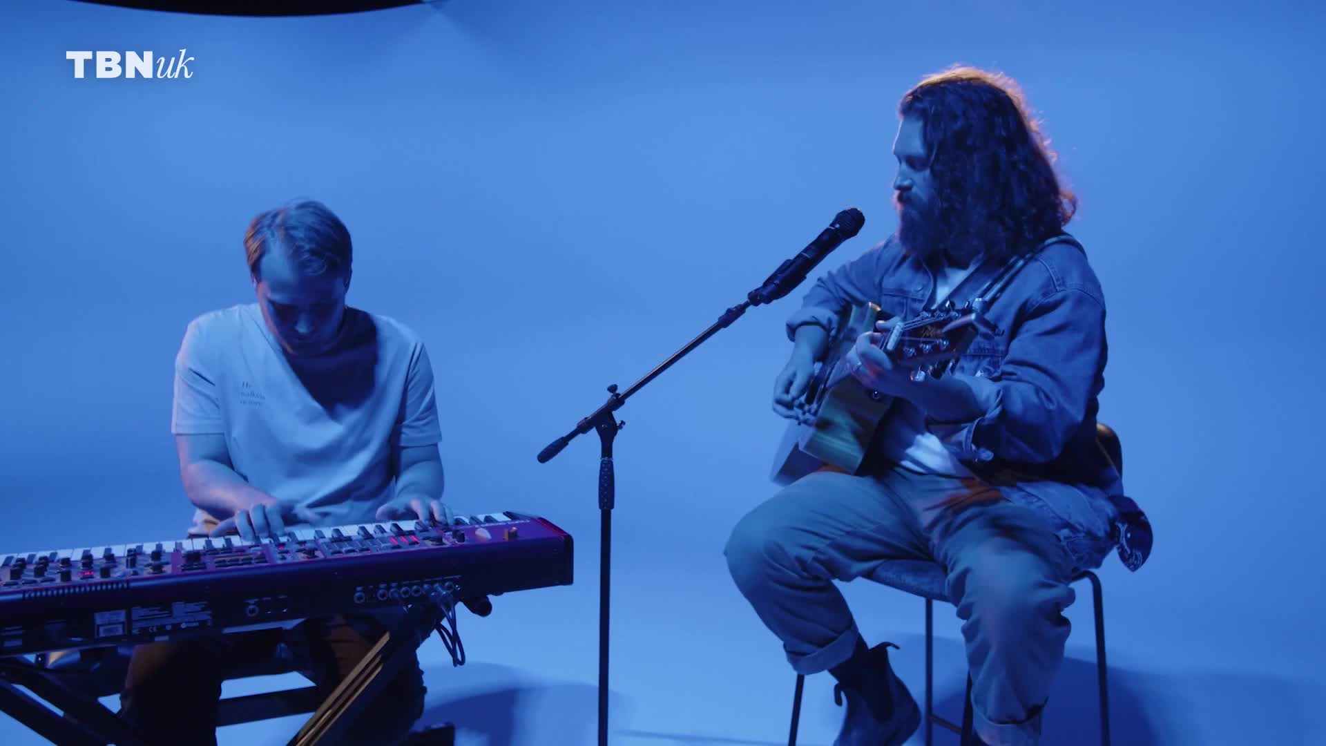 A man with long hair strums a guitar, while another plays a keyboard. The studio is bathed in blue light, the TBN UK logo visible in the corner.
