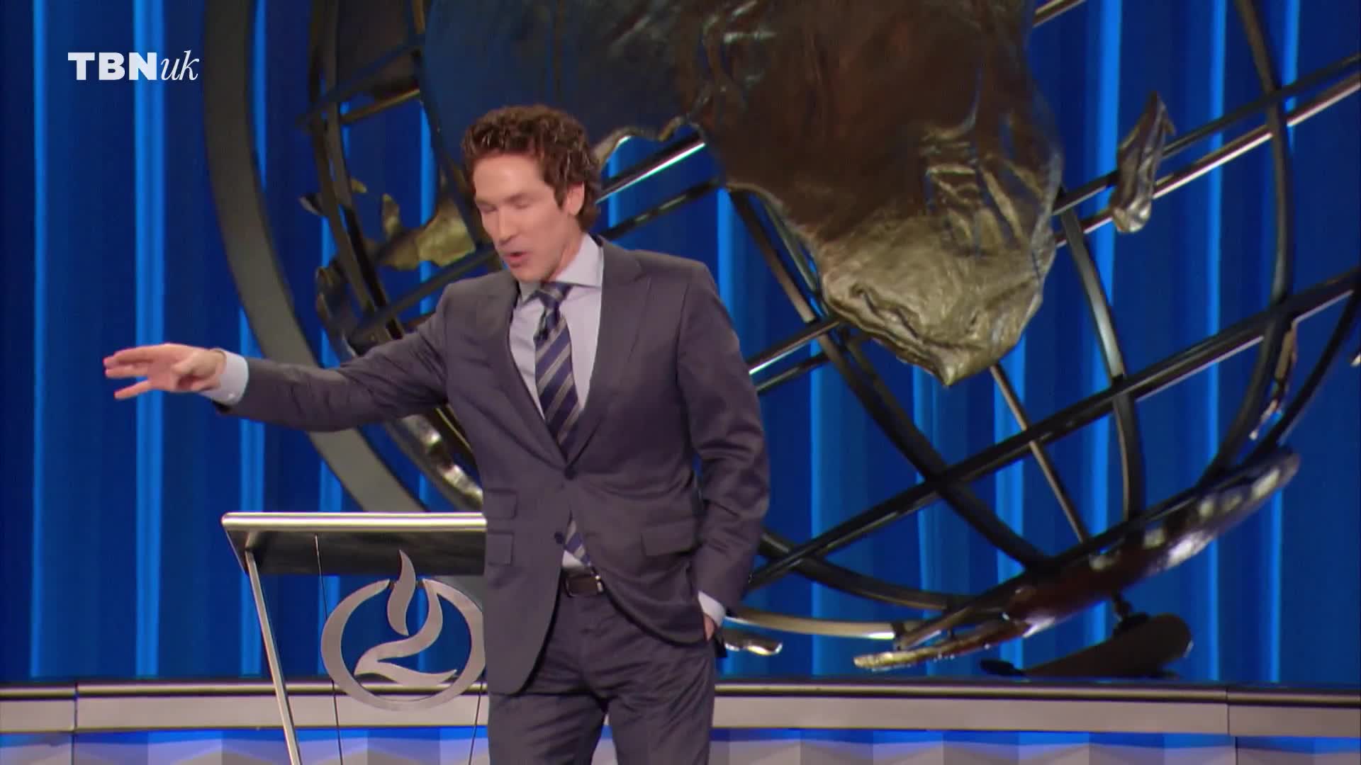 The man in the suit gestures with his right hand, speaking from behind a podium. Behind him, a metallic globe sits against a backdrop of blue vertical panels.
