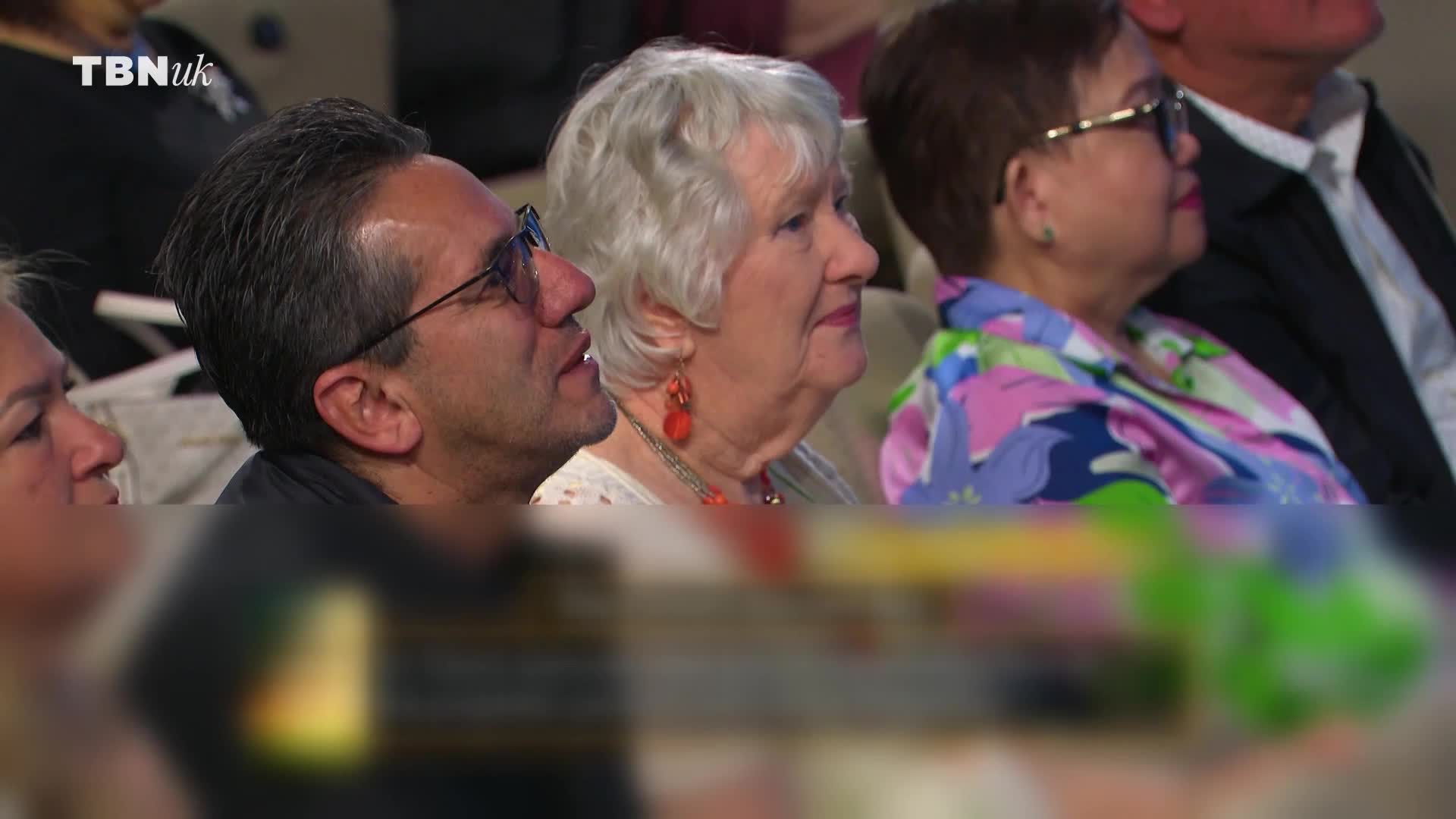 A man in glasses looks upward, a slight smile on his face. Beside him, a woman with white hair and red earrings listens intently to something happening on the TBN UK broadcast.
