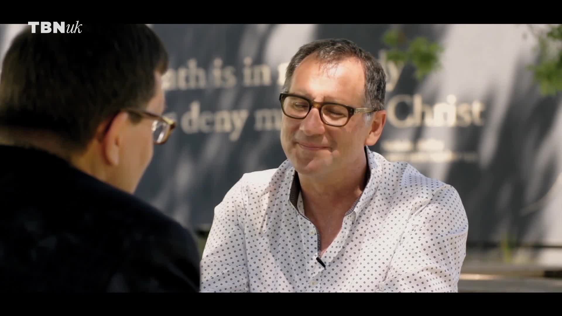 A man in a polka-dot shirt smiles, his eyes crinkling behind his glasses. He's sitting outside, facing another man who's mostly out of frame.
