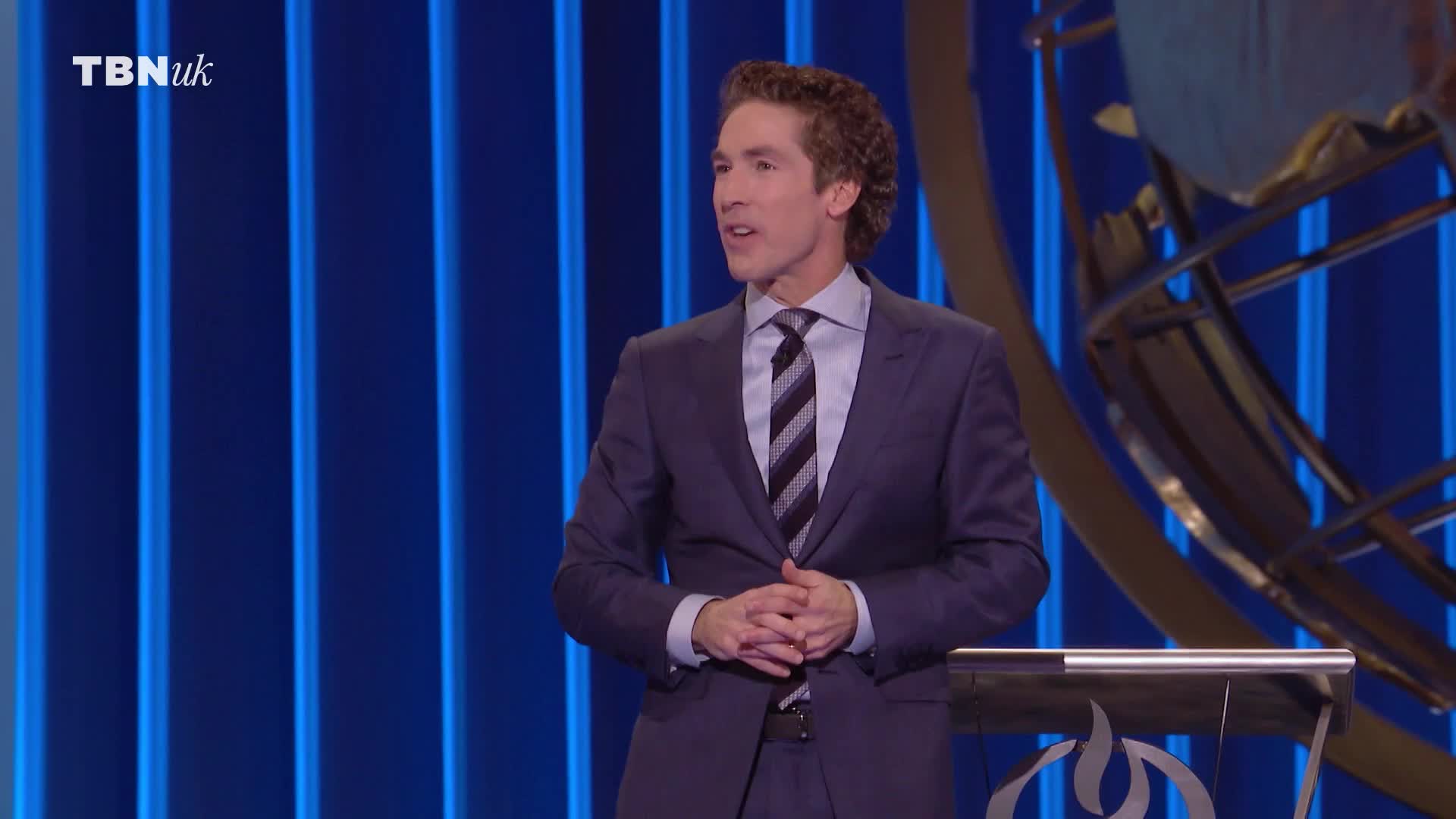 He's standing at a podium, hands clasped, speaking on TBN UK. The background is a dark blue with vertical light strips.
