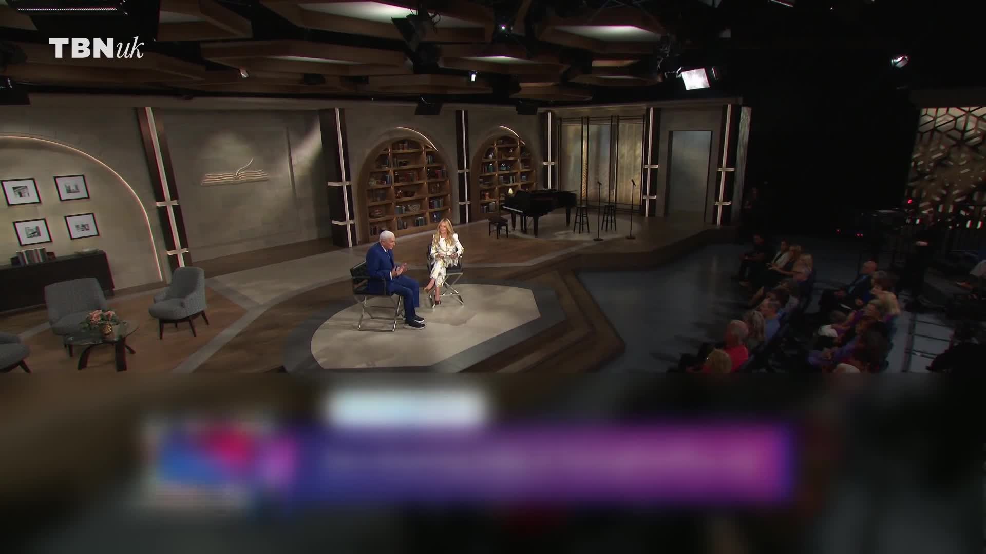 A man in a blue suit and a woman in a white dress sit facing each other on a stage, probably for a TBN UK interview. The audience is seated to the right, and a grand piano sits in the background.
