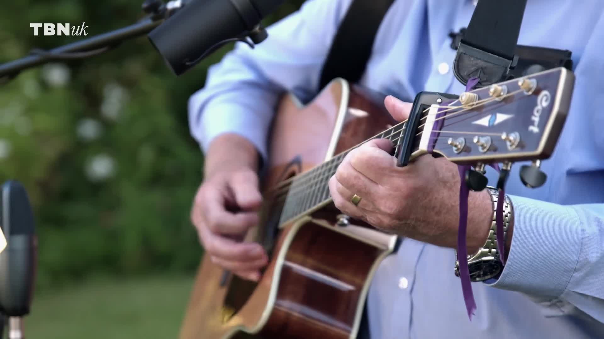 The man in the blue shirt strums a wooden acoustic guitar, his fingers moving across the strings. A microphone stands nearby, and the TBN UK logo is visible in the corner.
