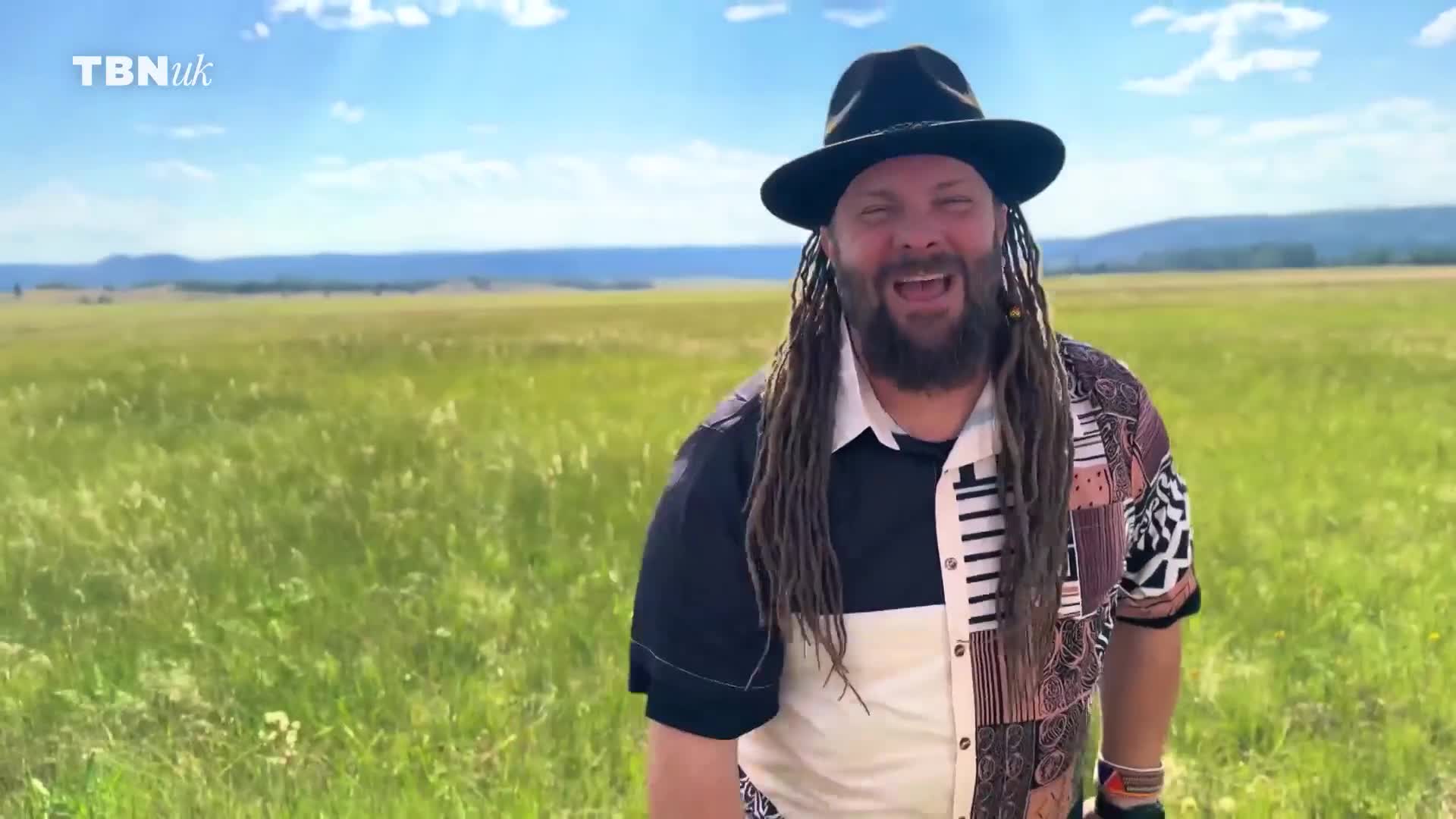 A man with dreadlocks and a hat stands in a field, smiling and talking. The TBN UK logo sits in the top left corner of the frame.
