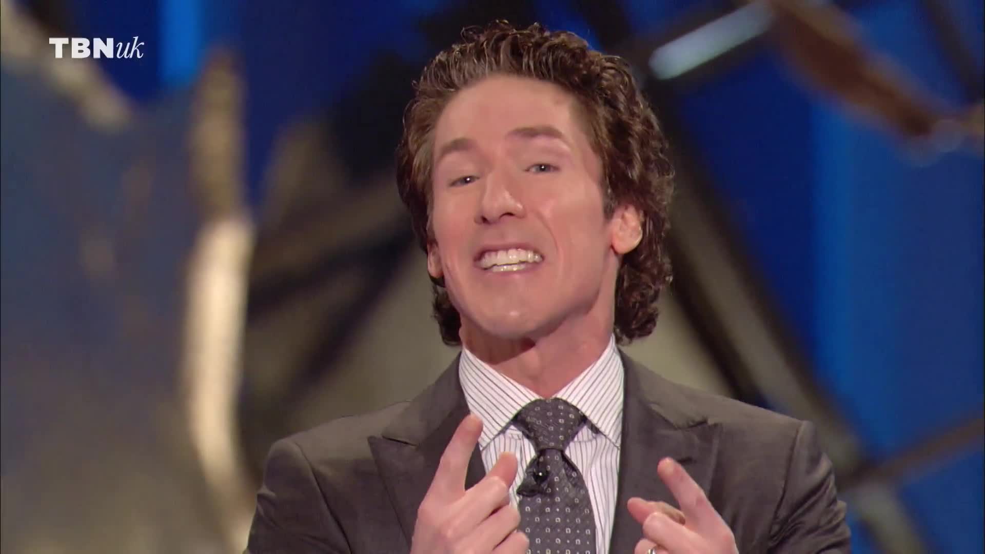 A man in a suit gestures with both hands, his fingers pointing upwards. He's speaking on a TBN UK broadcast, his expression animated.
