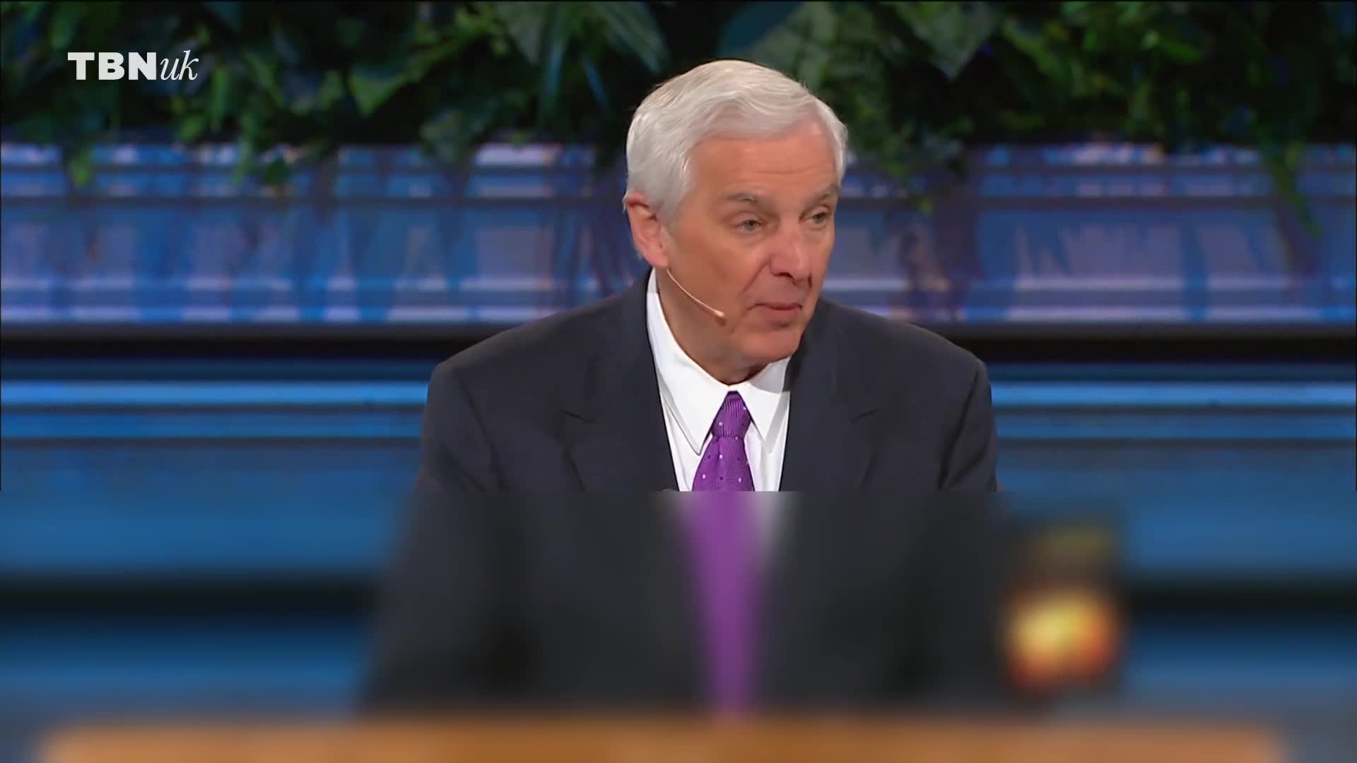 A man in a suit is speaking, a microphone clipped to his lapel. The TBN UK logo is visible in the corner, and the backdrop is a soft blue.
