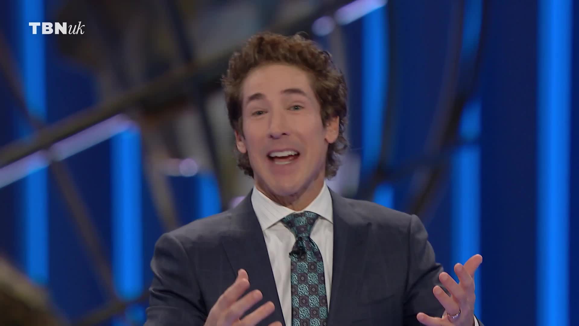A man in a suit, seen on TBN UK, is speaking, his hands open and gesturing. His expression is animated, his mouth open as if mid-sentence.
