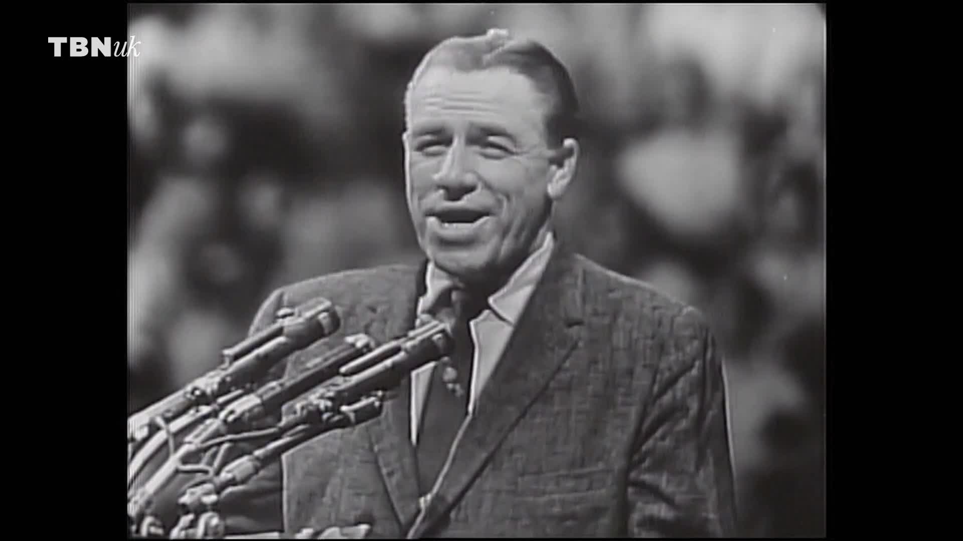 A man in a suit, speaking into a cluster of microphones, is the focus. The TBN UK logo is visible in the corner, and a crowd is blurred in the background.
