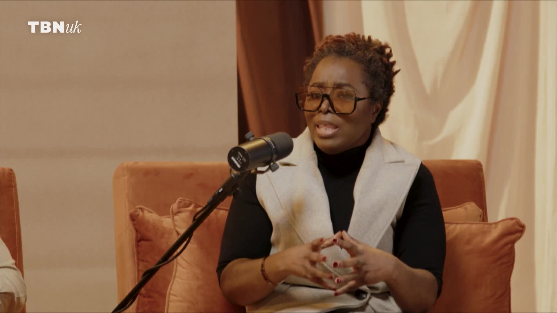 A woman in glasses gestures with her hands while speaking into a microphone. She's seated on a plush, orange chair, part of a set for a TBN UK broadcast.
