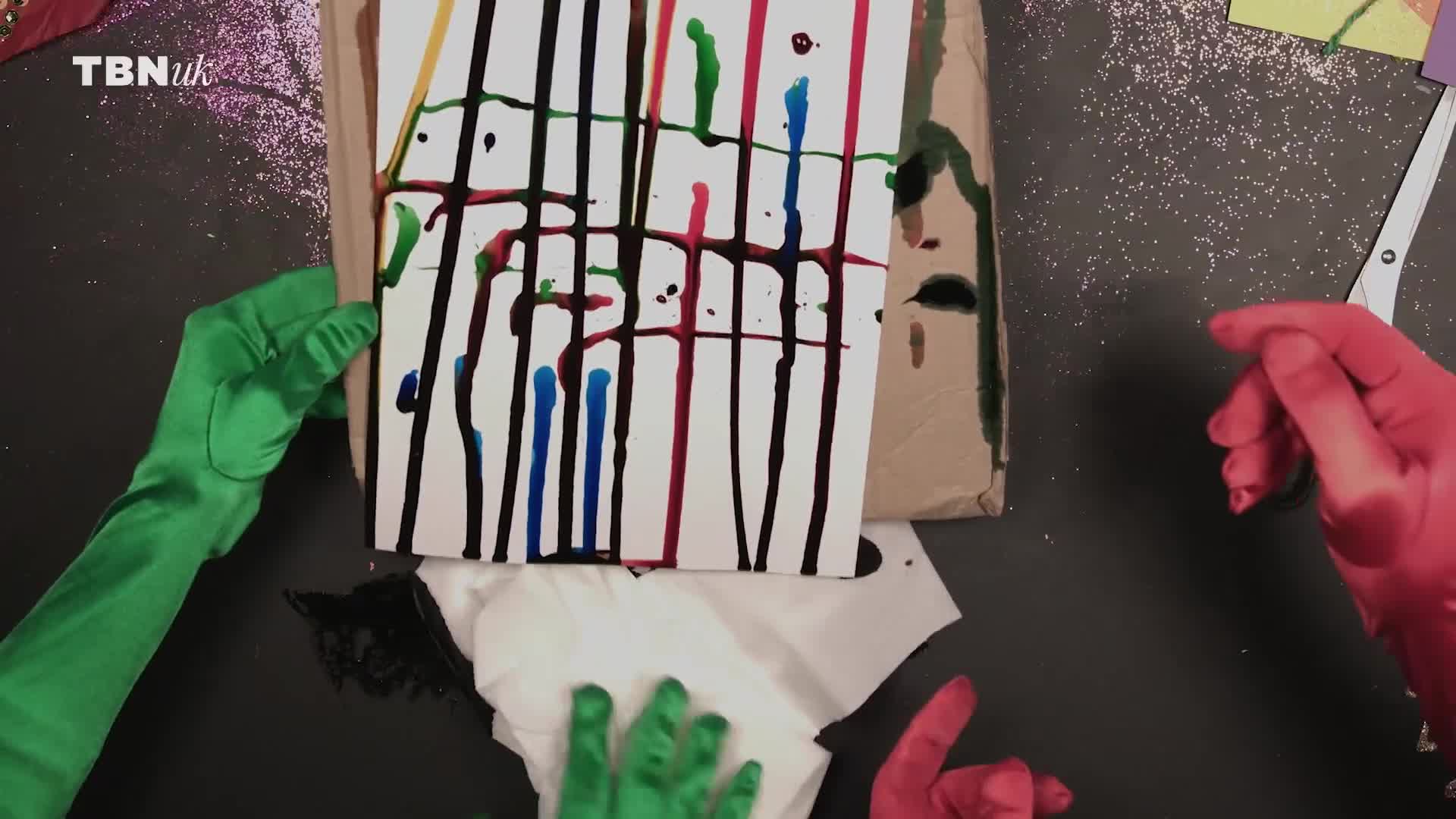 A sheet of paper, striped with drips of paint, is held up by a gloved hand. Another hand, also gloved, dabs at the paint with a tissue.
