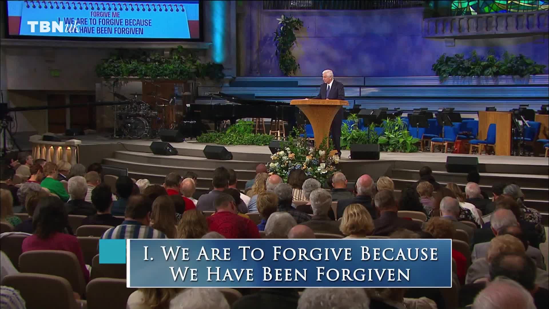 A man in a dark suit stands at a podium, addressing a seated audience. Above him, the TBN UK logo is visible, and a screen displays the words "We are to forgive because we have been forgiven."

