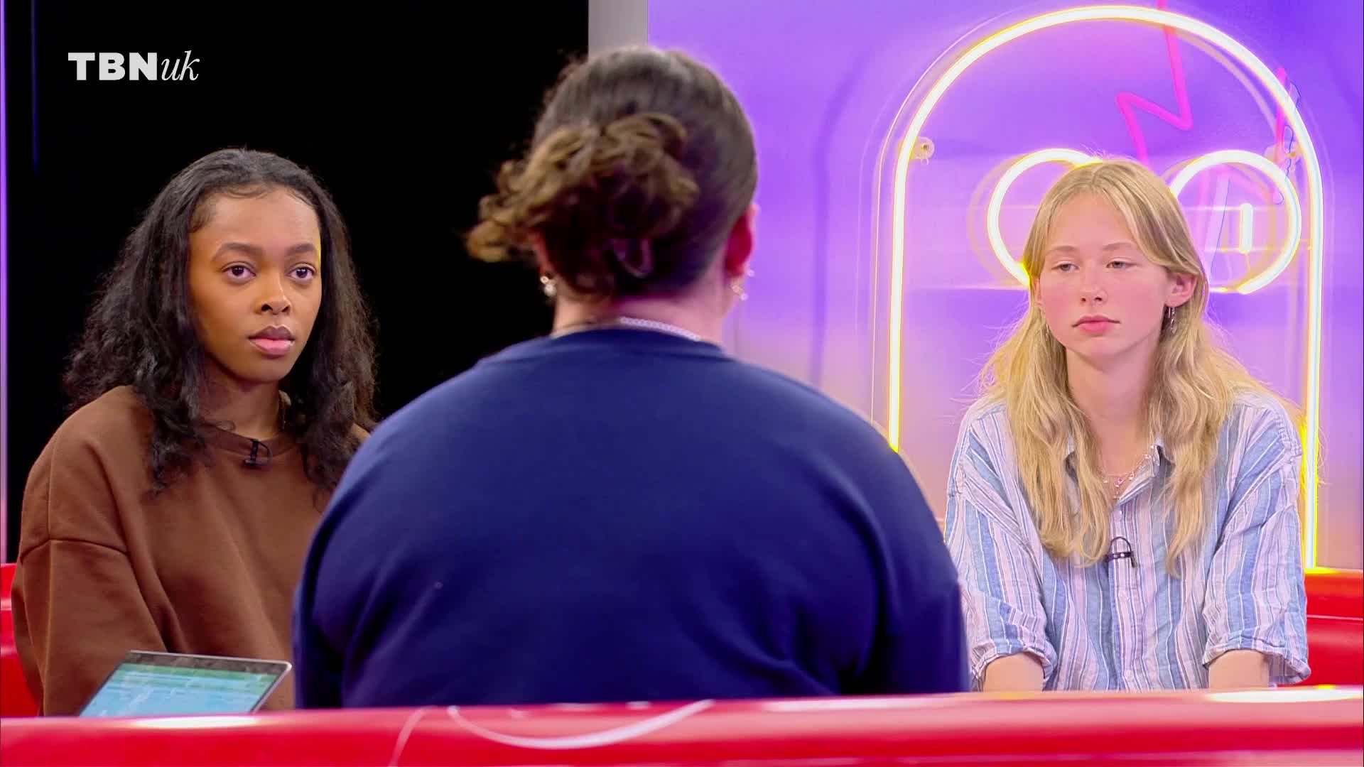 Three people are seated in a studio, the back of one's head facing the camera. Two young women sit on either side, both looking intently at the person in front of them.
