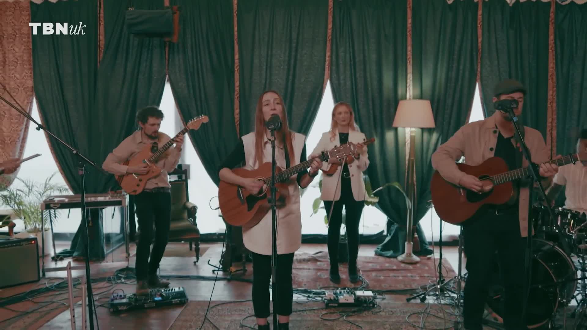 A band is playing in a studio, with a woman in the center singing and strumming an acoustic guitar. The TBN UK logo is visible in the top corner.

