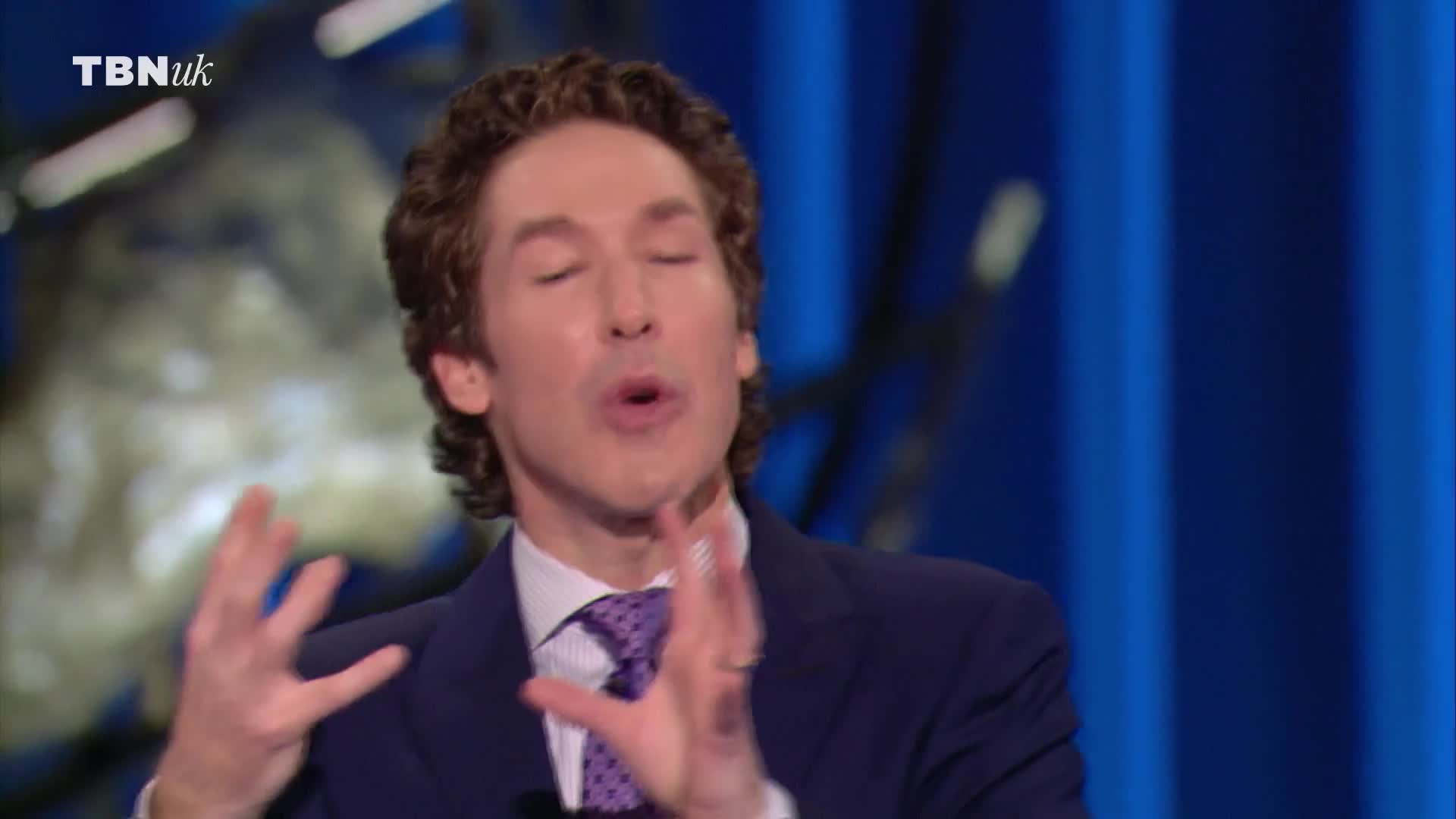He's speaking, his hands raised in a gesture, and his mouth is open. The TBN UK logo is visible in the corner.
