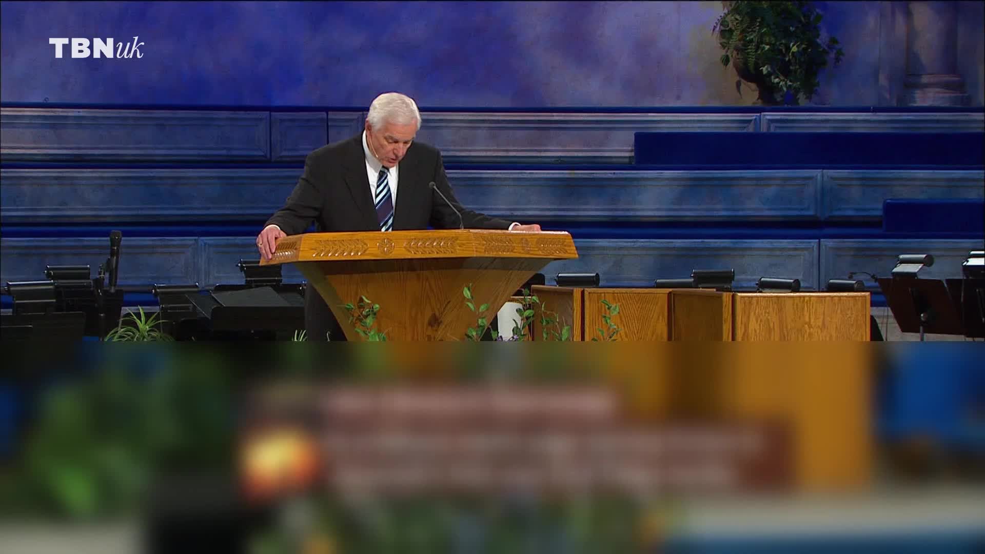 A man in a suit stands at a wooden lectern, reading from a script. The TBN UK logo is visible in the top left corner of the screen.
