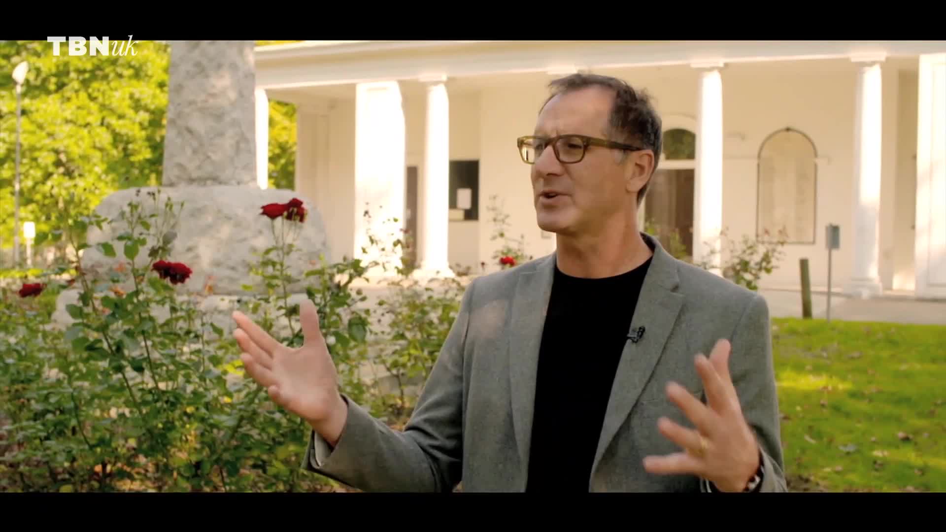 A man with glasses gestures with his hands, speaking in front of a white building and a rose bush. The TBN UK logo is visible in the top left corner.
