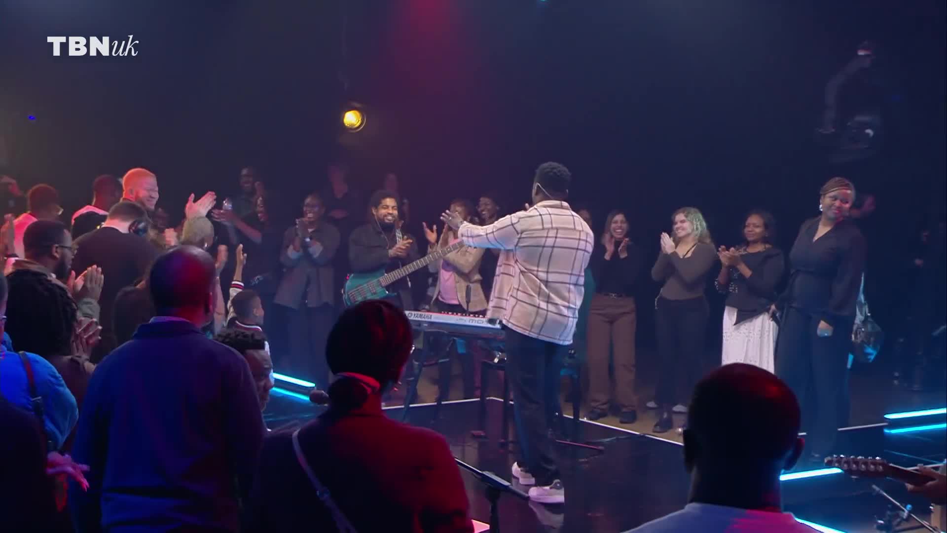 On a stage lit by neon, a man in a plaid shirt gestures to a crowd clapping and smiling. The TBN UK logo is visible as the band and choir stand ready.
