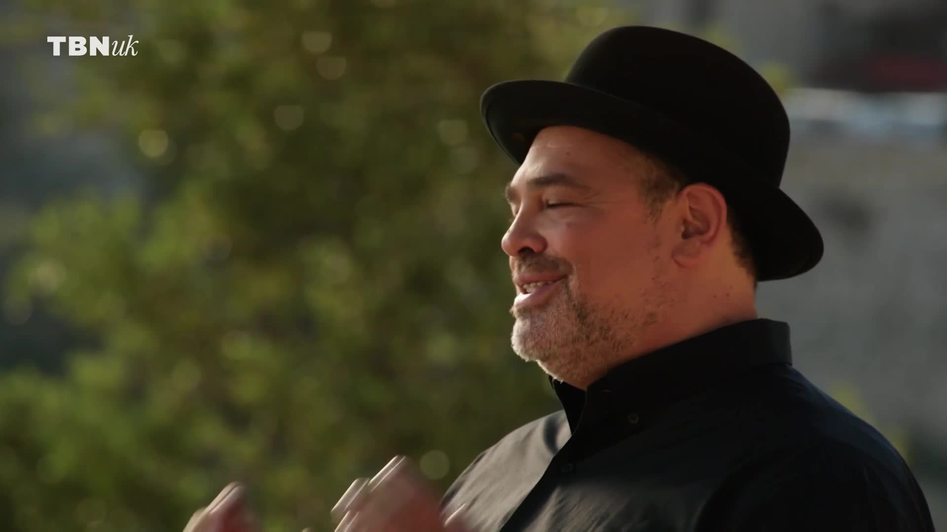 A man in a black hat and shirt smiles, his eyes closed as he speaks. The TBN UK logo is visible in the corner.

