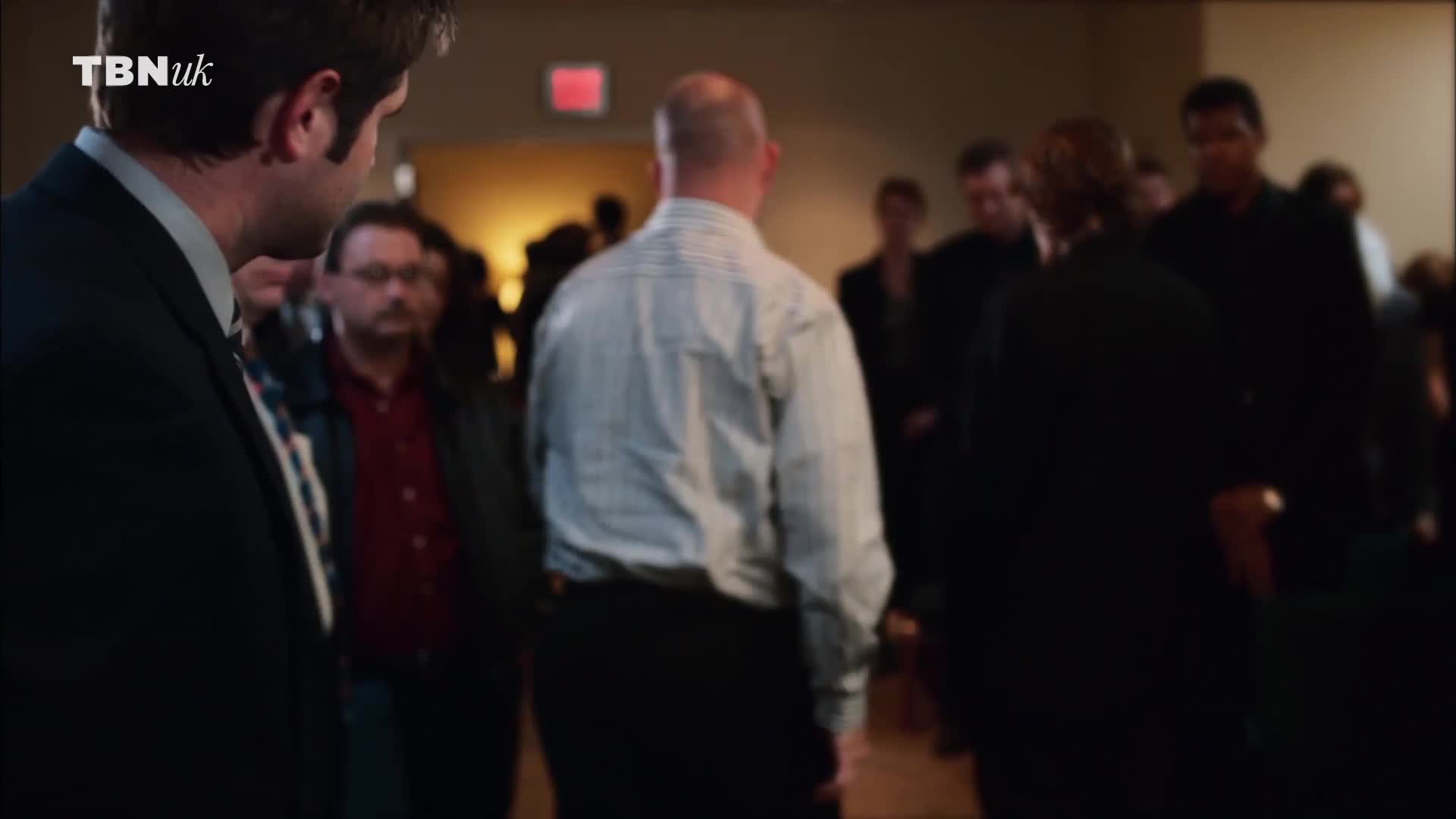 A man in a striped shirt walks forward, his bald head catching the light. Behind him, a group of people in dark suits and jackets mill about, the TBN UK logo visible in the corner.
