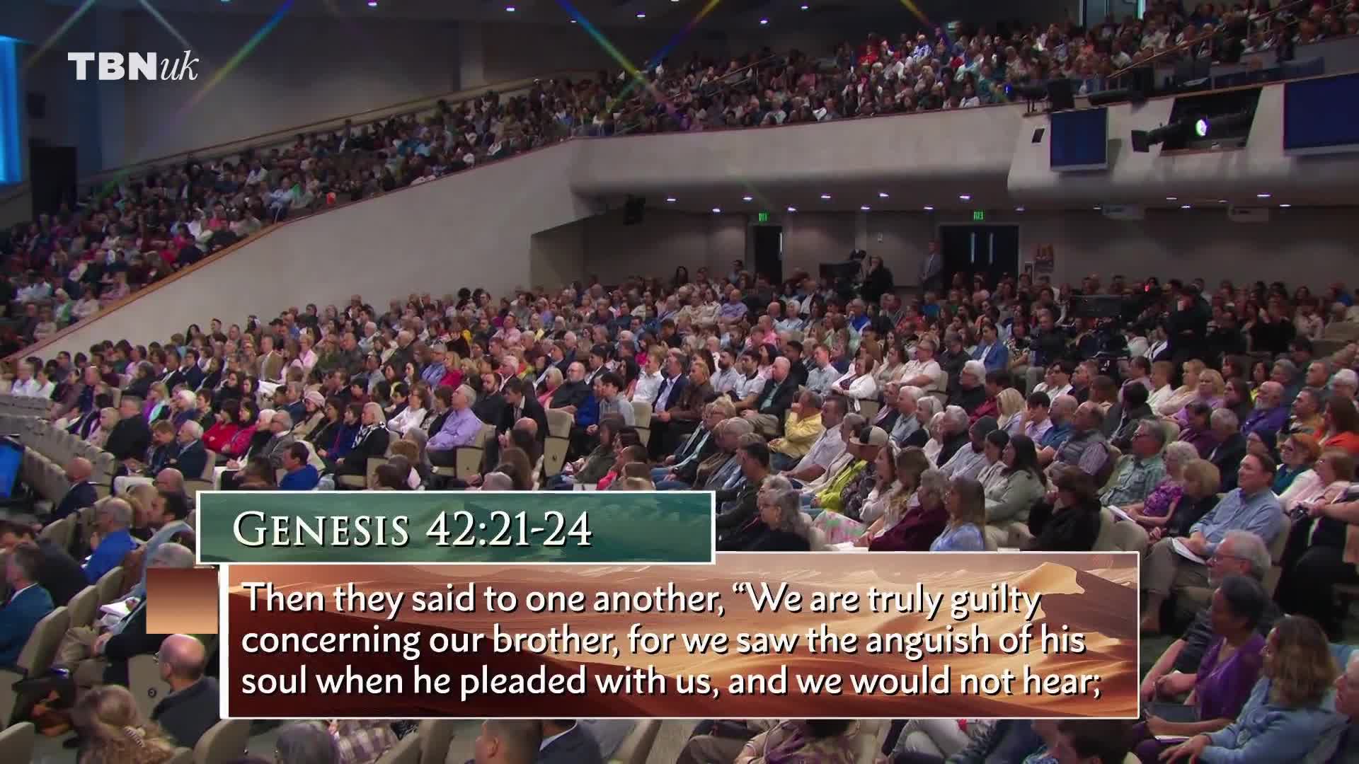 A large crowd sits in tiered seating, listening intently. A passage from Genesis is displayed on screen, likely part of a broadcast from TBN UK.
