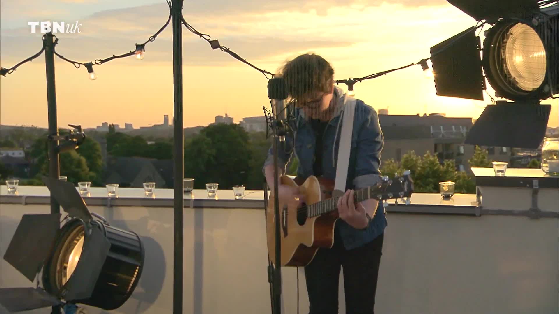 On a rooftop in the United Kingdom, a musician strums an acoustic guitar, lit by spotlights and string lights as the sun sets. The TBN UK logo is visible as he performs, with a cityscape visible in the background.
