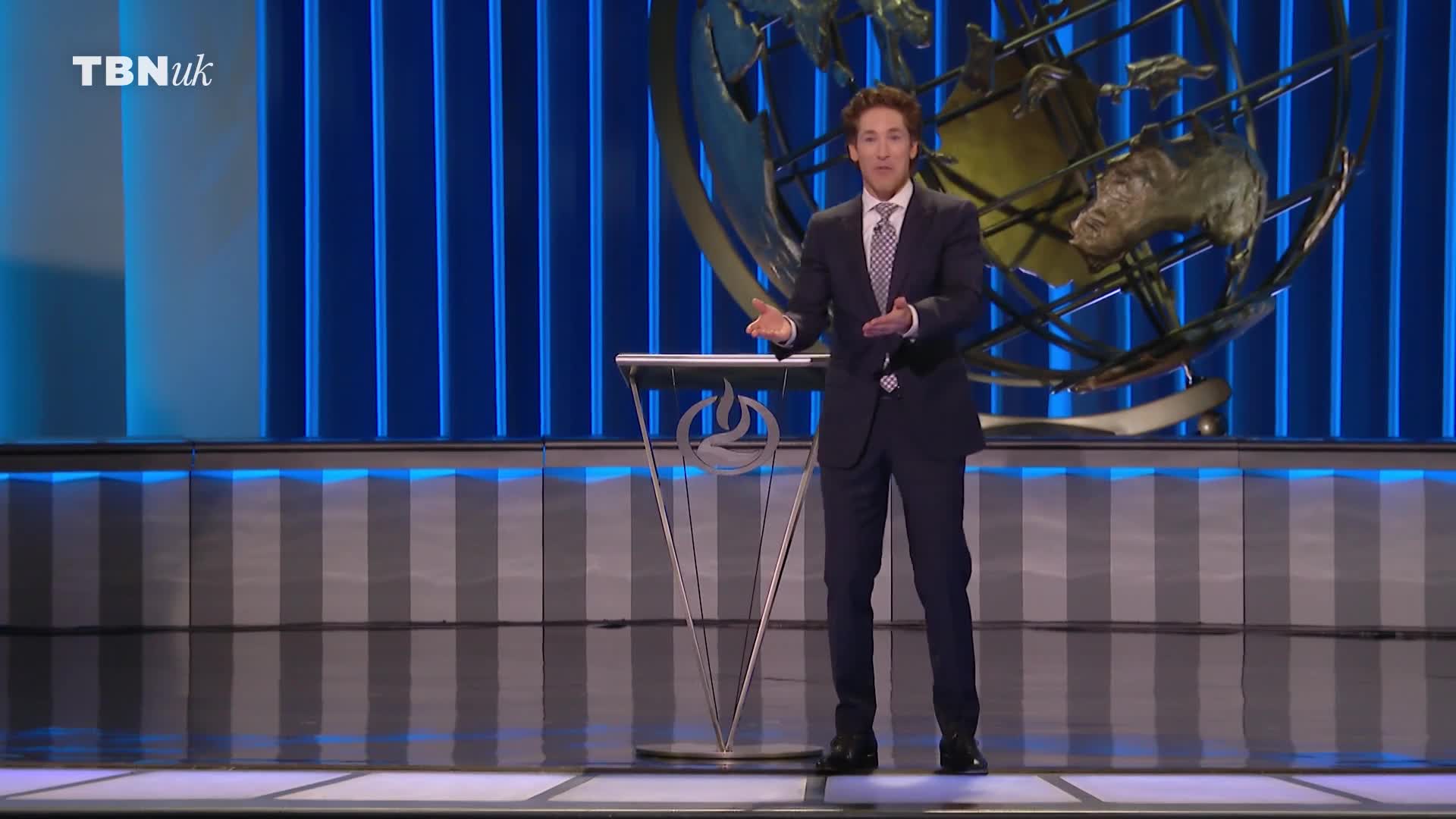 A man in a dark suit stands center stage, gesturing with both hands. The backdrop features a large, ornate globe and the TBN UK logo.
