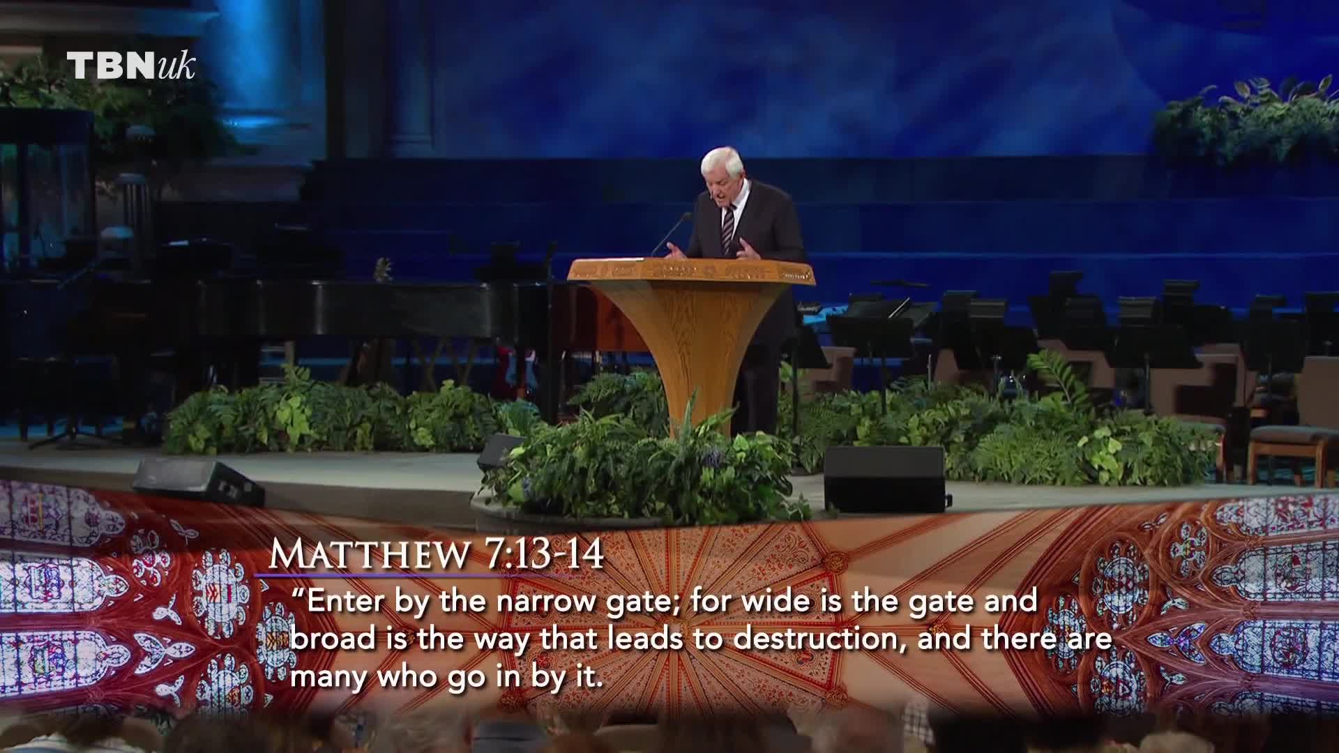 A man in a suit stands at a wooden lectern, speaking into a microphone on the TBN UK stage. Lush greenery surrounds the platform, and behind him, an orchestra is set up.
