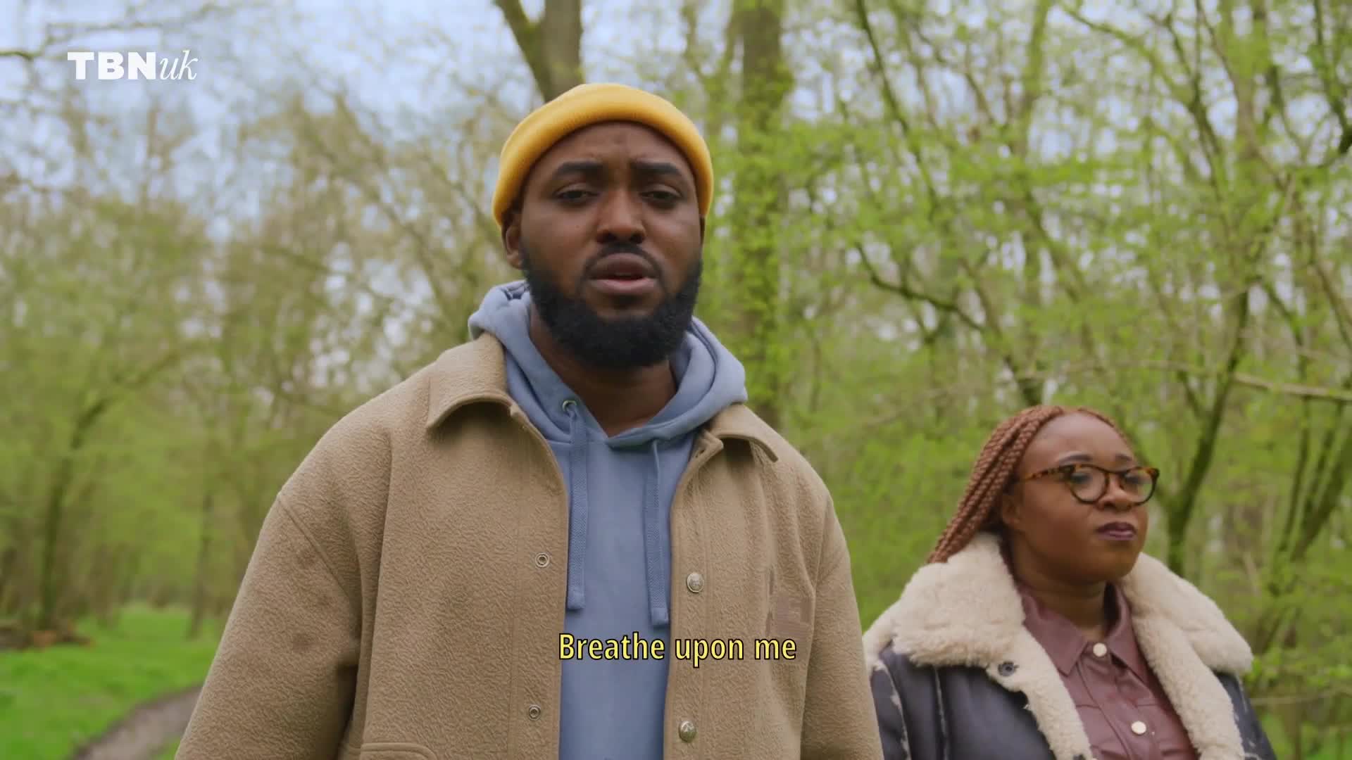 A man in a yellow beanie and tan jacket looks up, speaking. Beside him, a woman in glasses and a leather jacket listens intently in the UK, on TBN UK.

