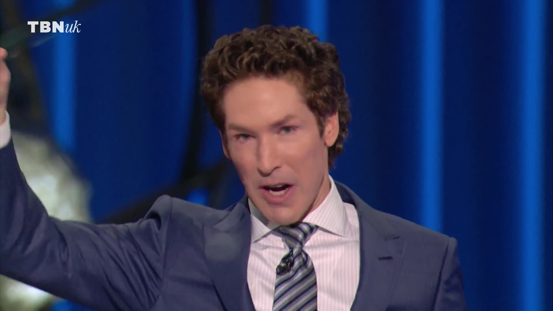 A man in a suit, speaking with his mouth open, gestures with his right arm raised. The TBN UK logo is visible in the top left corner.
