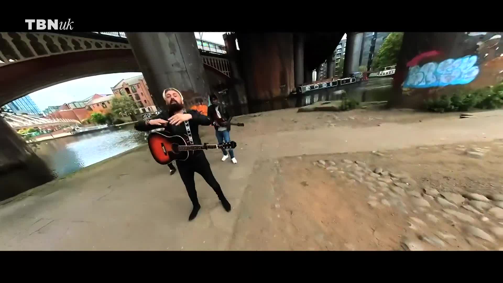 Underneath a bridge in the UK, a man with a beard strums an acoustic guitar, flanked by other musicians. The band performs near a graffiti-covered wall beside a canal, with the TBN UK logo visible in the corner.
