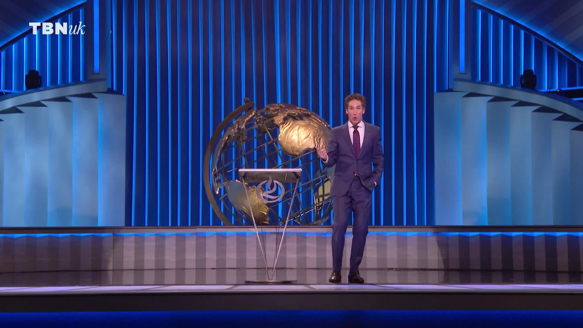 The man in the suit gestures with his right hand, pacing the stage. Behind him, a globe rests on a stand, and the TBN UK logo is visible in the corner.
