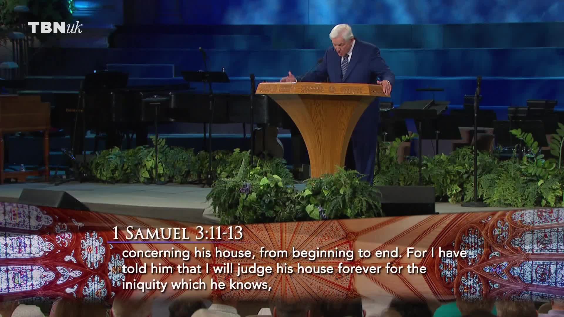 A man in a blue suit stands at a wooden lectern, gesturing with his hands. The TBN UK logo is visible, and text from 1 Samuel is displayed on the screen below.
