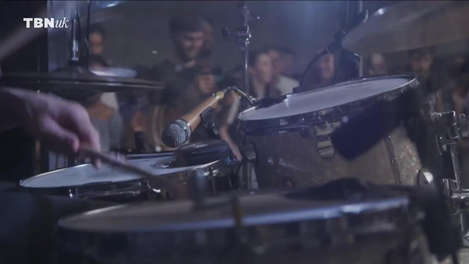 A drummer's hand raises a stick, poised to strike a snare drum. Behind the kit, a crowd of faces is barely visible, watching the performance on TBN UK.
