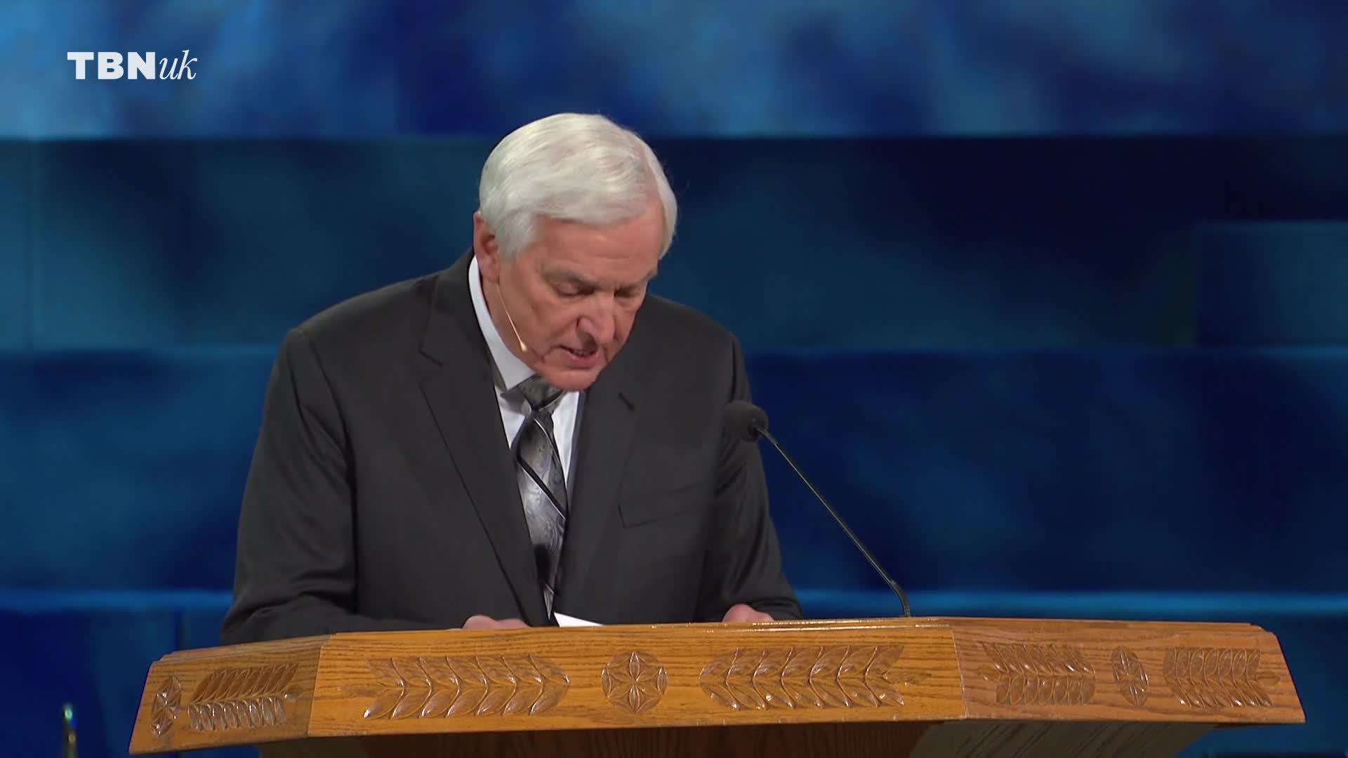 A man in a dark suit stands behind a wooden lectern, his gaze fixed downwards as he reads. The TBN UK logo is visible in the corner.
