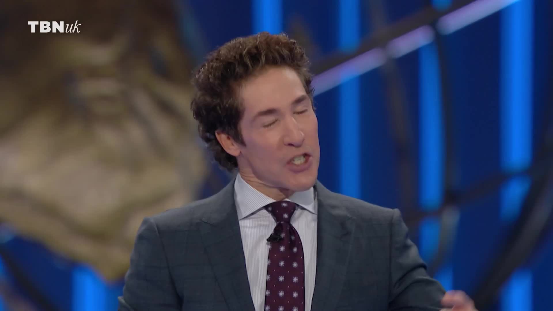 A man in a dark suit and patterned tie is speaking, his mouth open as he addresses the audience. The TBN UK logo is visible in the top left corner.
