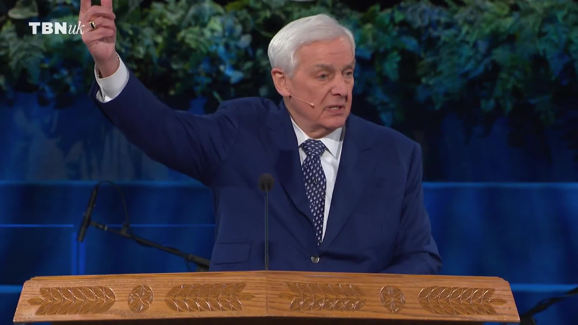 The man in the blue suit gestures with his right hand, his index finger pointing upwards. He's speaking from behind a wooden lectern, the TBN UK logo visible in the corner.
