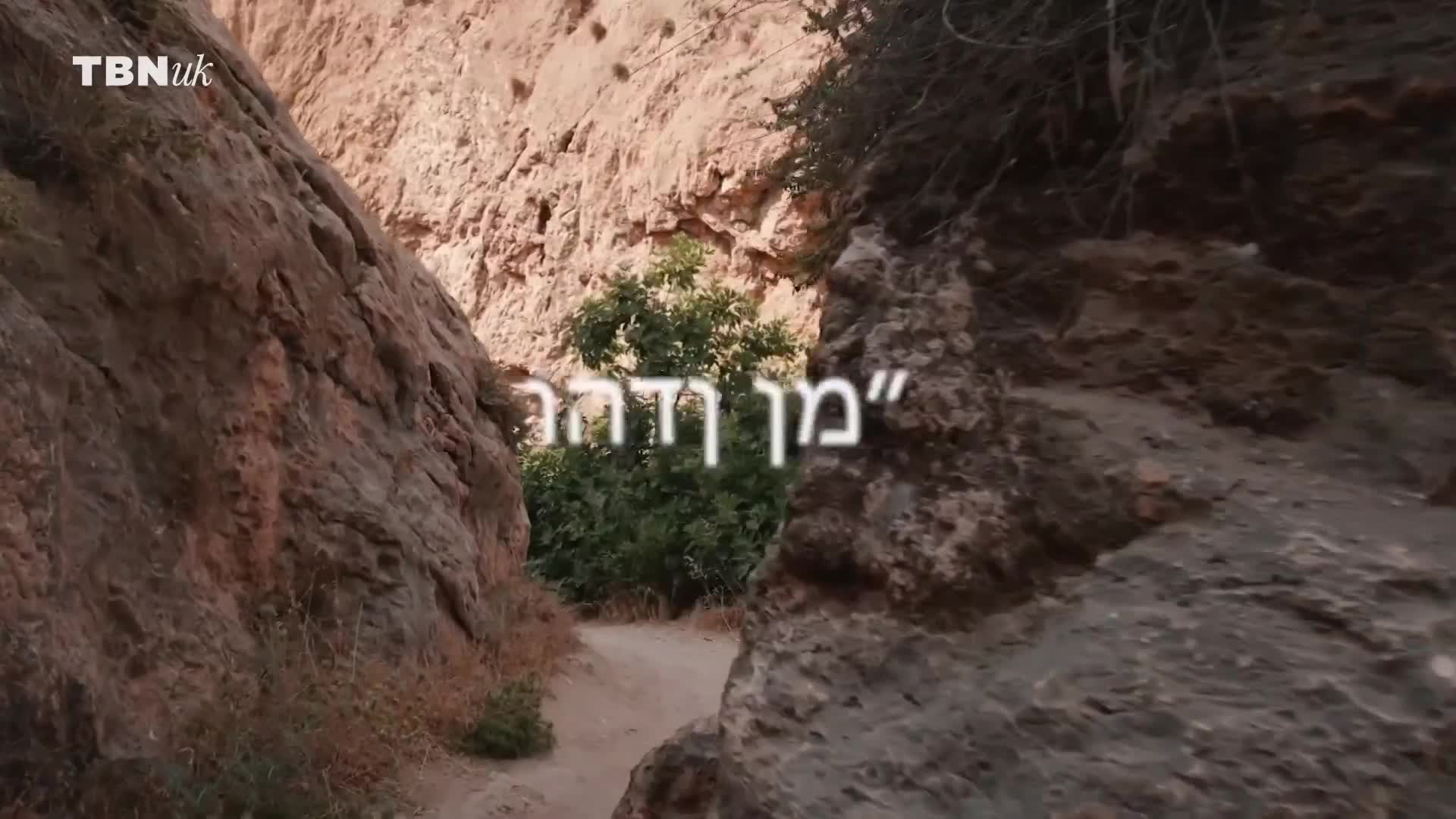 The camera peers through a narrow passage between two rock faces, framing a patch of green foliage at the end. The TBN UK logo is in the corner, and the Hebrew text floats in the center.
