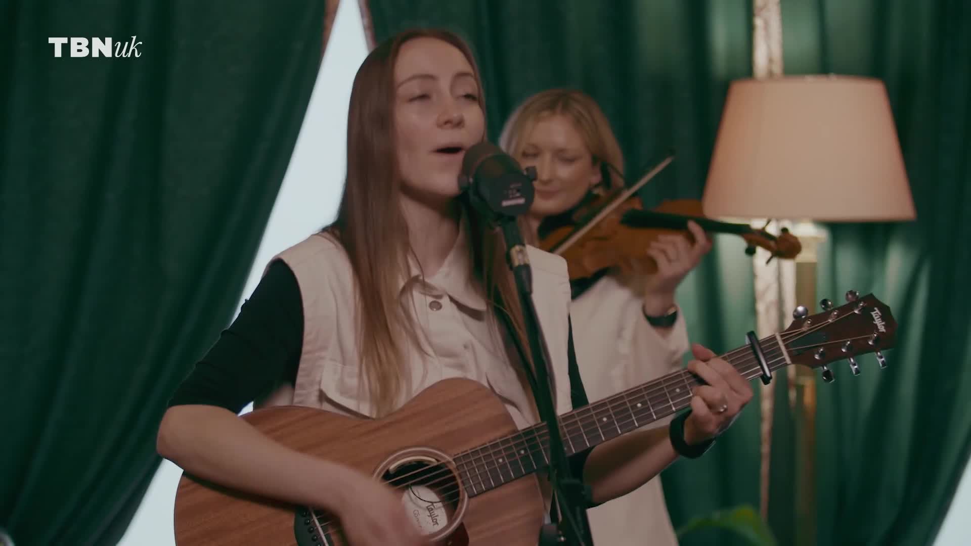 A woman strums a guitar and sings into a microphone while another plays the violin behind her. The TBN UK logo is visible in the corner.
