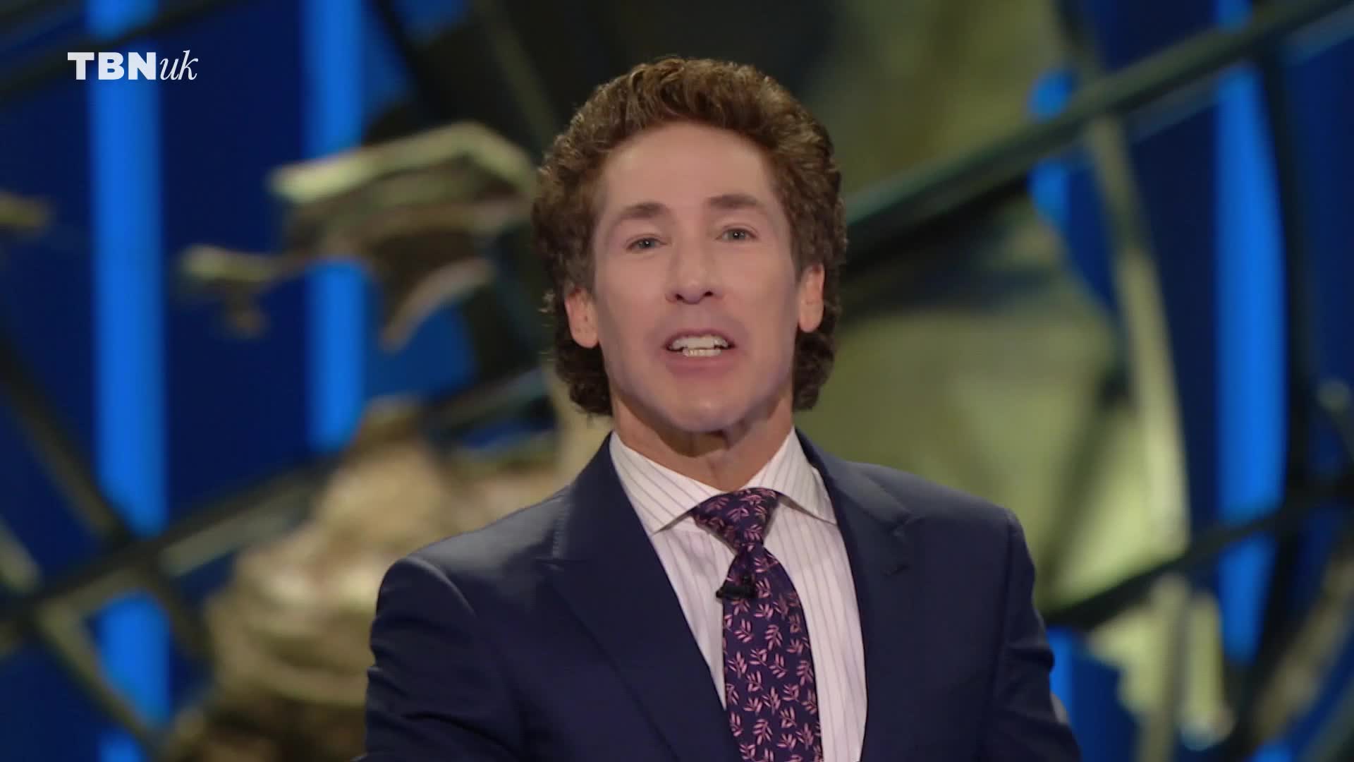 He's speaking, his mouth moving as he addresses the camera. The TBN UK logo is visible in the corner, and the background is a blend of blue and abstract shapes.
