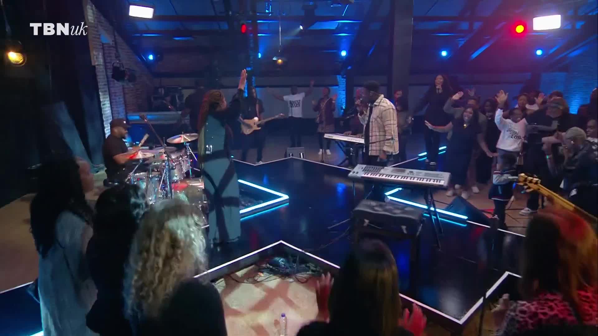 A band is playing on a stage shaped like a star, bathed in blue and red lights. The crowd in front of the stage is clapping and raising their hands while a singer in a plaid shirt belts out a song.
