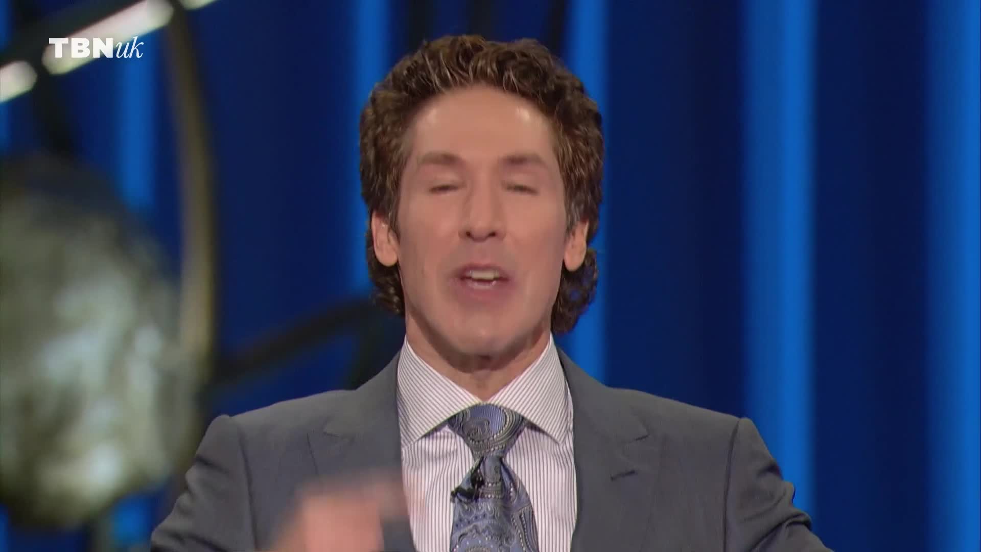 The man in the suit is speaking, his eyes closed as he addresses the audience on TBN UK. His hands are moving, and the background is a solid blue.
