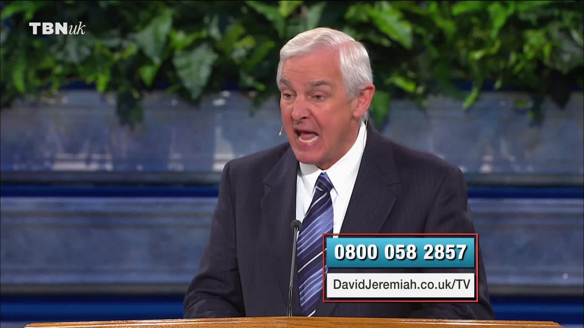 A man in a dark suit is speaking, his mouth open wide as he gestures. The TBN UK logo is visible, and a phone number is displayed on the screen.
