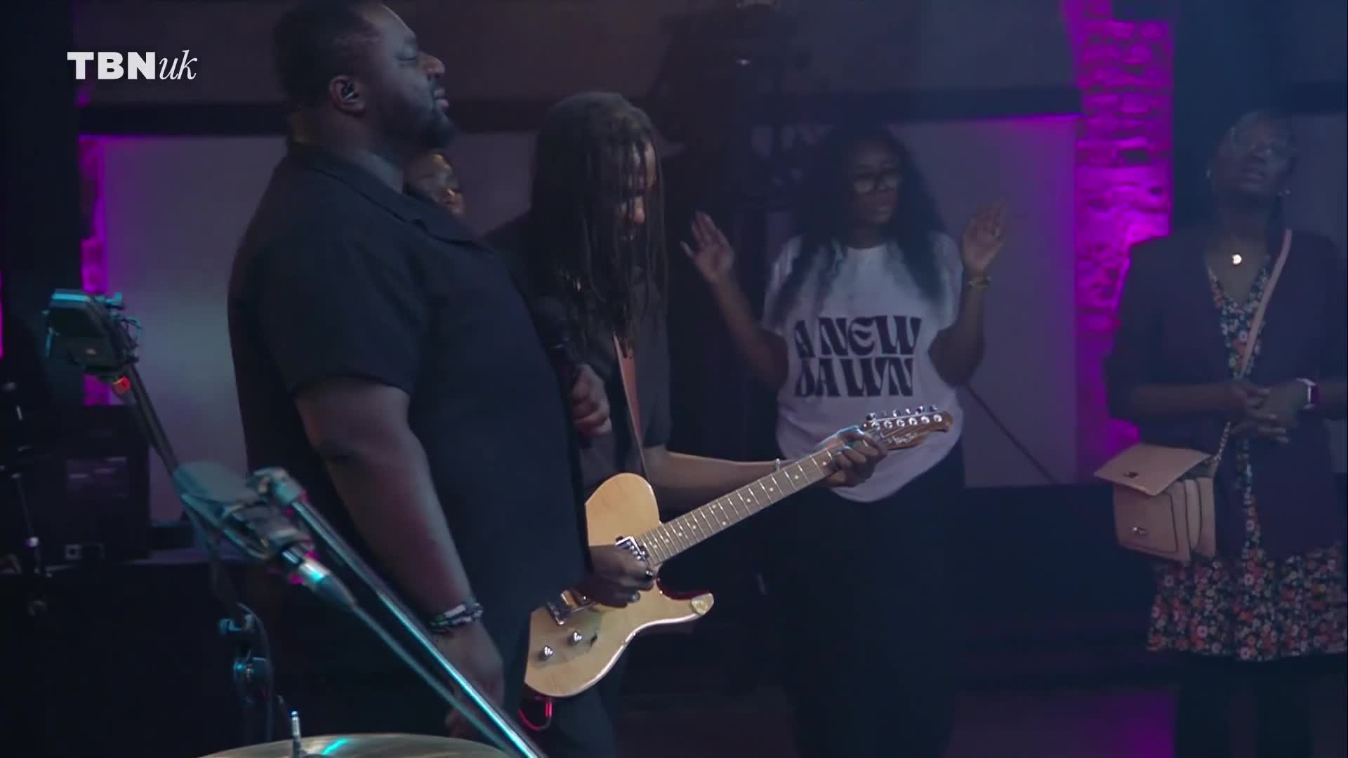 A guitarist strums a light-colored electric guitar, while others around him raise their hands. The scene is bathed in purple and white light, likely from a TBN UK broadcast in the United Kingdom.
