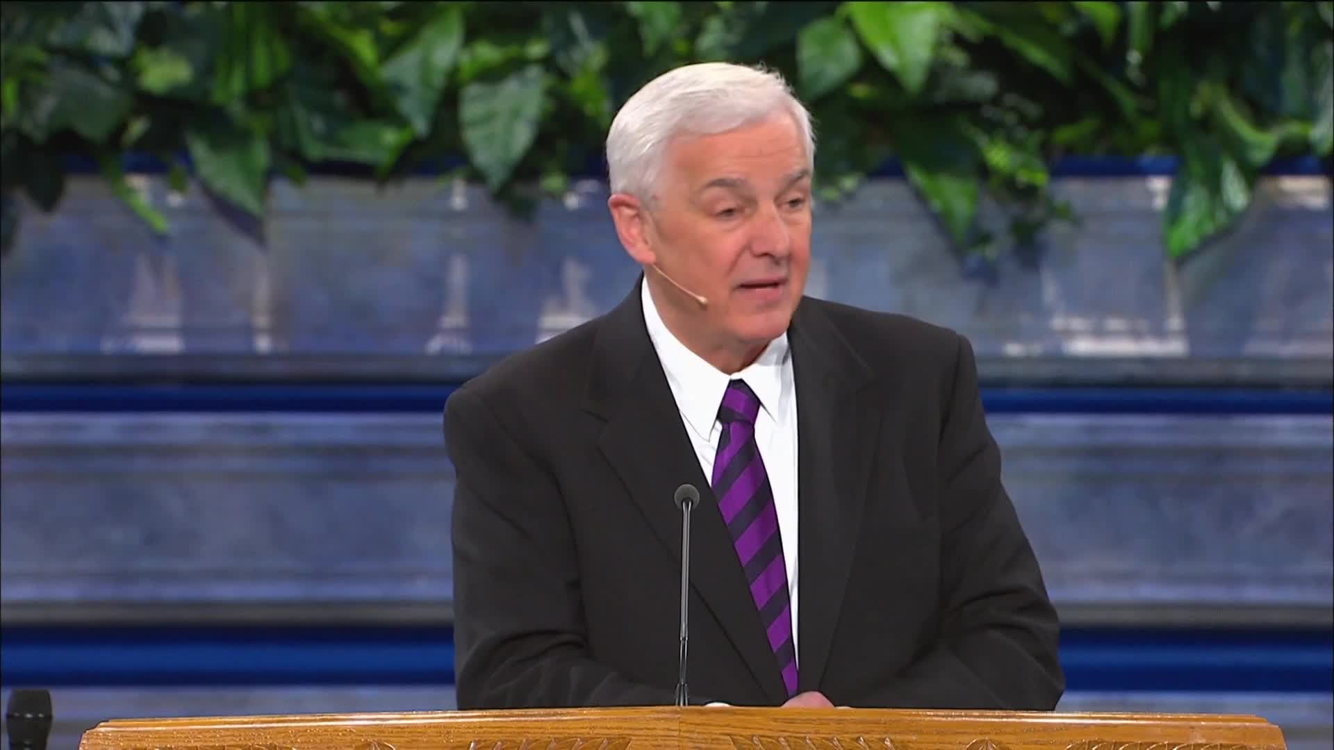 A man in a suit with a purple tie is speaking into a microphone. He stands behind a wooden podium, with a backdrop of greenery and a blue-toned wall.
