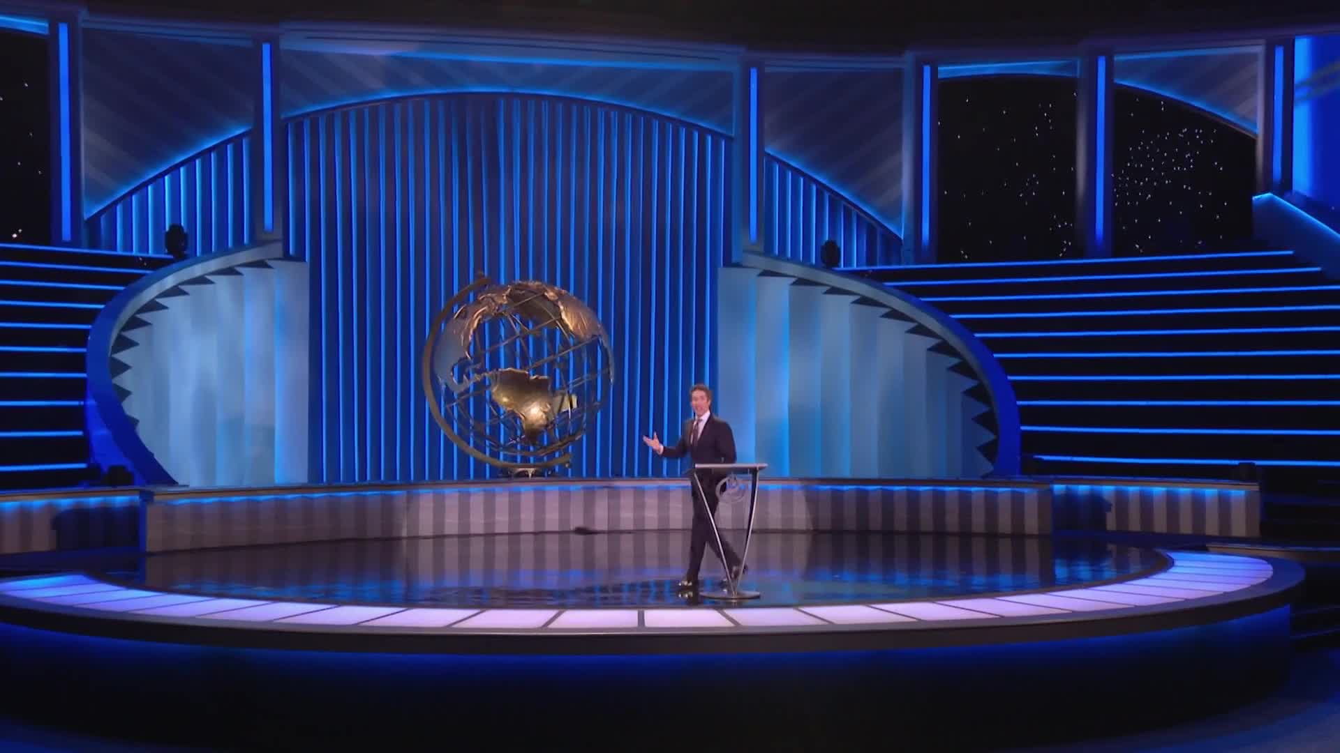 A man in a suit stands at a podium on a stage, gesturing to the audience.  Behind him, a large globe sculpture is centered against a backdrop of blue lights and steps, likely for a TBN UK broadcast.
