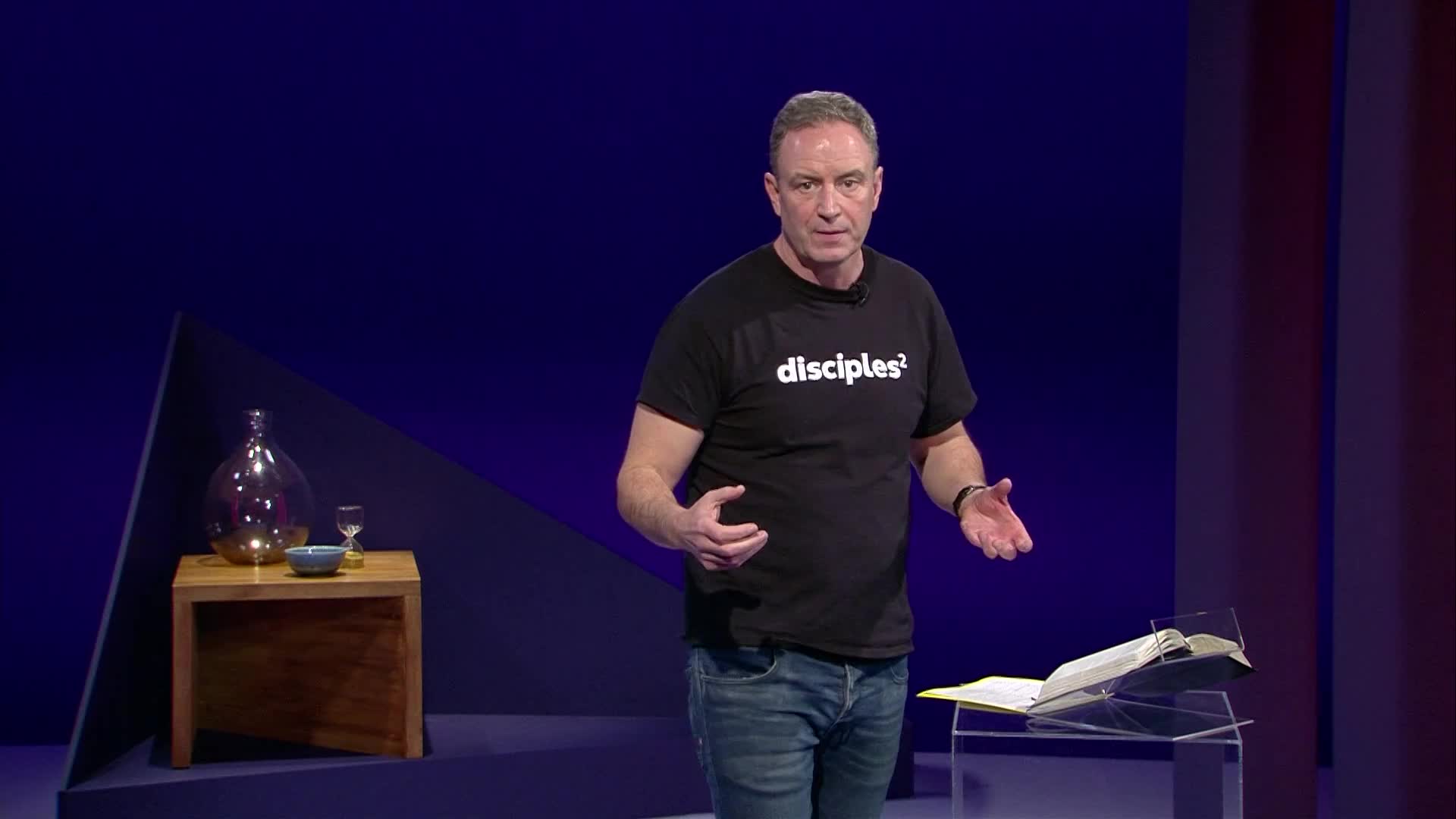 A man in a black t-shirt and jeans gestures with his hands as he speaks. An open book rests on a clear stand beside him, and a small table holds a glass bottle and other items.
