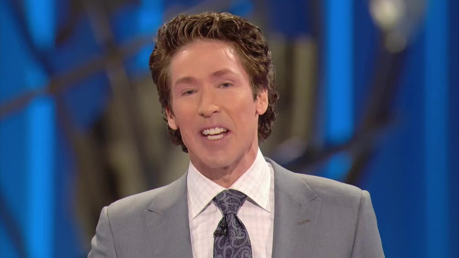 He's speaking directly to the camera, a light grey suit framing his face. Behind him, the background is a soft, blurred blue.
