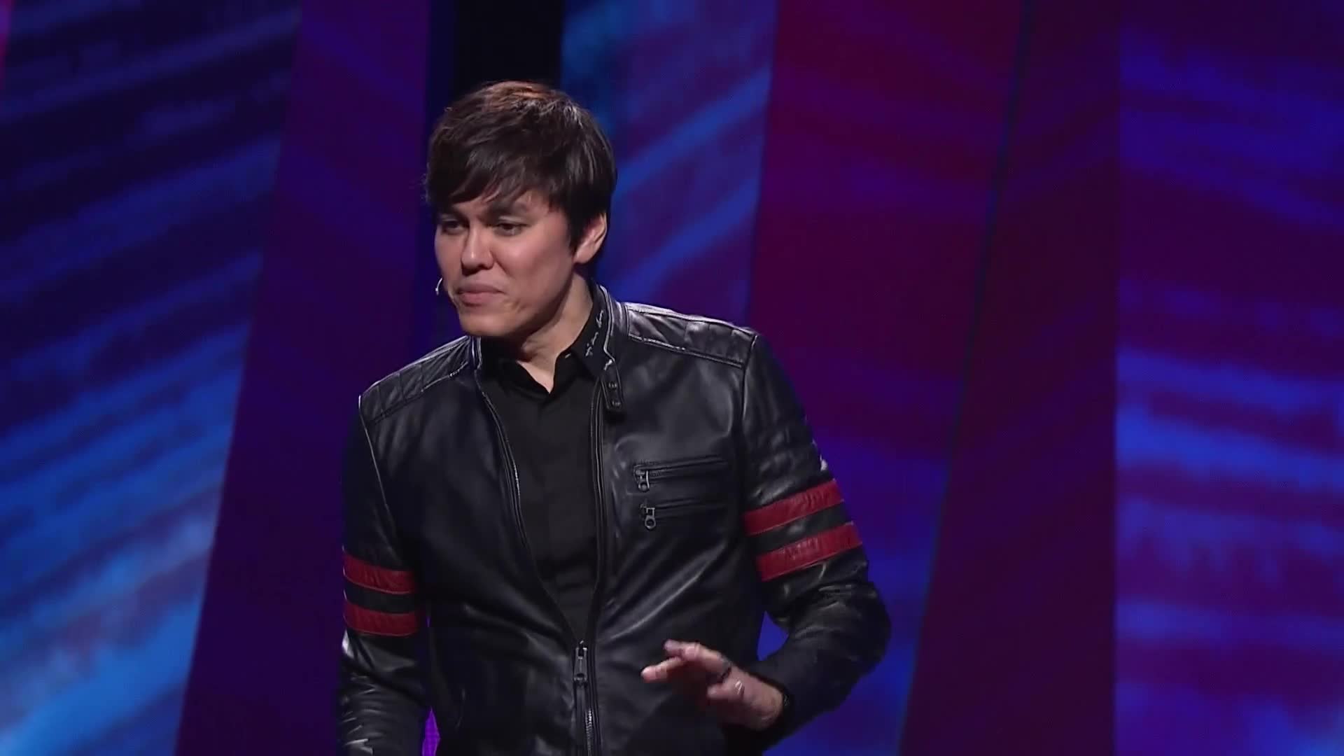 He's standing on a stage, gesturing with his right hand. His black leather jacket has red stripes on the sleeves, and he's speaking to the audience.
