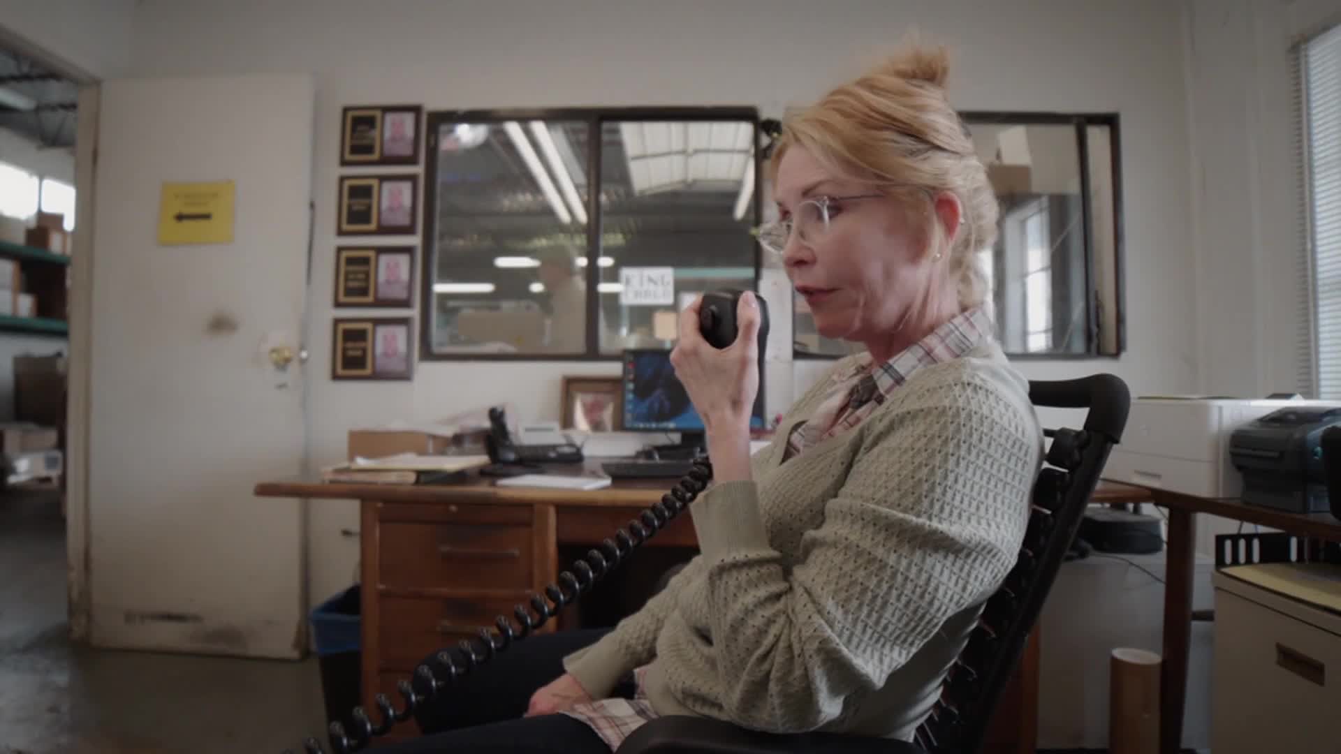 A woman with glasses sits in an office chair, speaking into a microphone. The coiled cord stretches across her lap, leading to a desk cluttered with papers and a computer.
