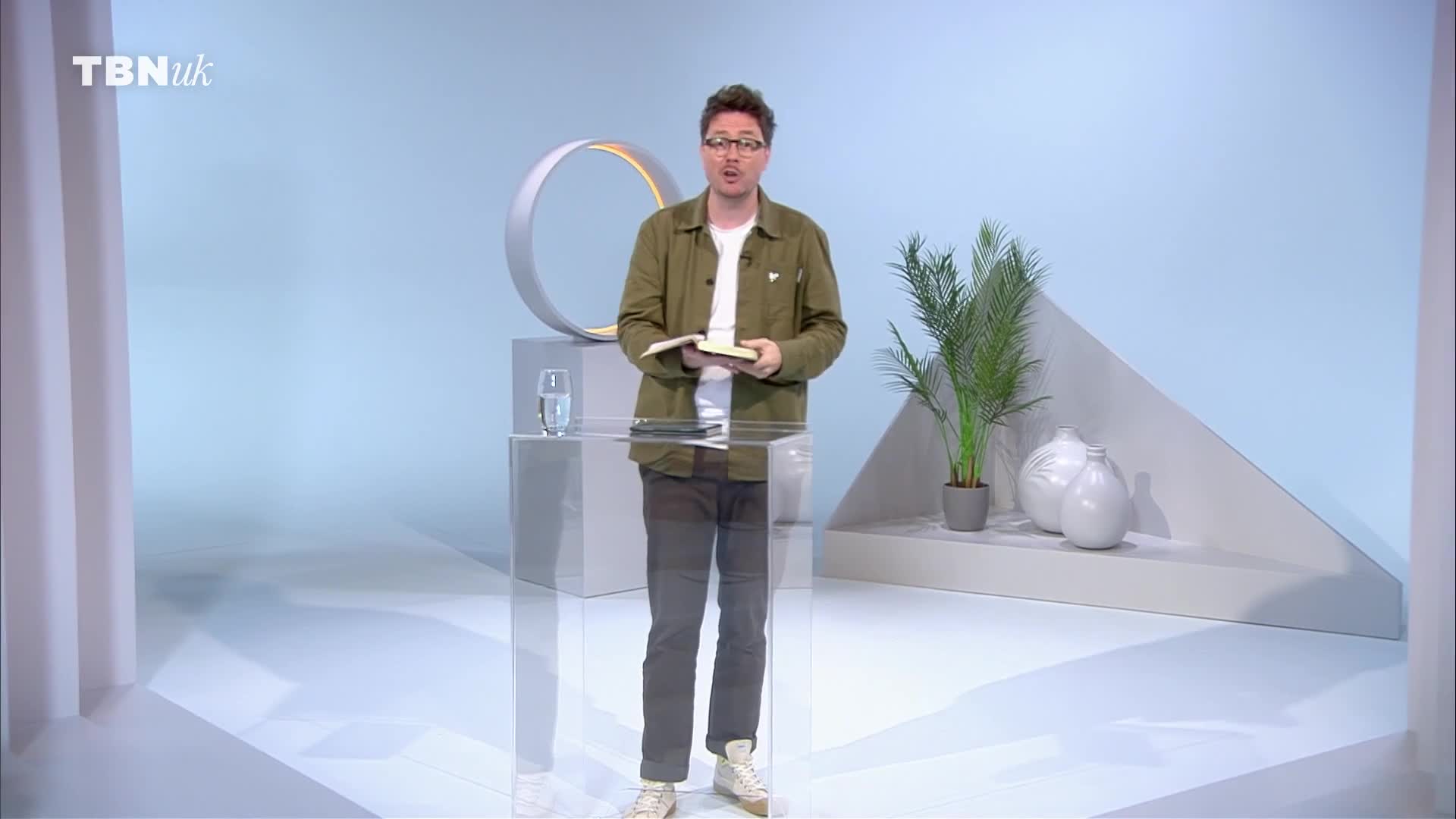 A man in a green jacket stands at a clear podium, holding a book, presumably on a TBN UK set. He's speaking to the camera, the backdrop a minimalist design with a plant and a decorative circle.
